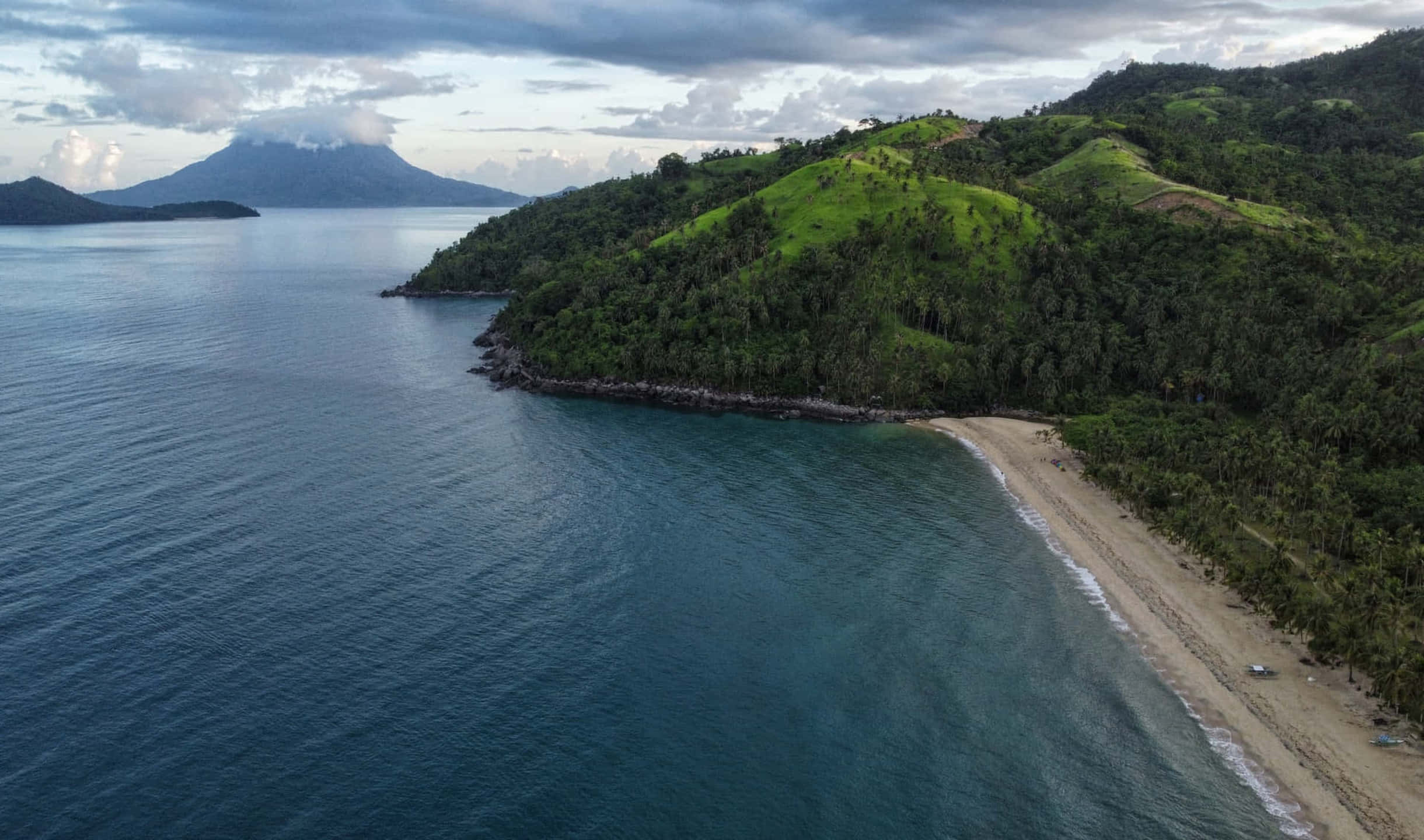 Philippines 桌布 壁紙 — 鳥瞰圖：一個隱蔽的熱帶海灘沿著翠綠山丘蜿蜒，遠處有一座火山，天空多雲。