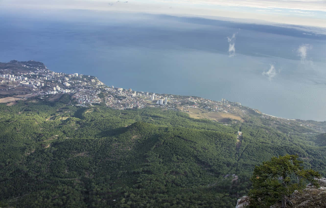島嶼 桌布 壁紙 — 鳥瞰圖：一座沿海城市坐落在鬱鬱蔥蔥的綠色山丘中，旁邊是廣闊的藍色大海，天空多雲。