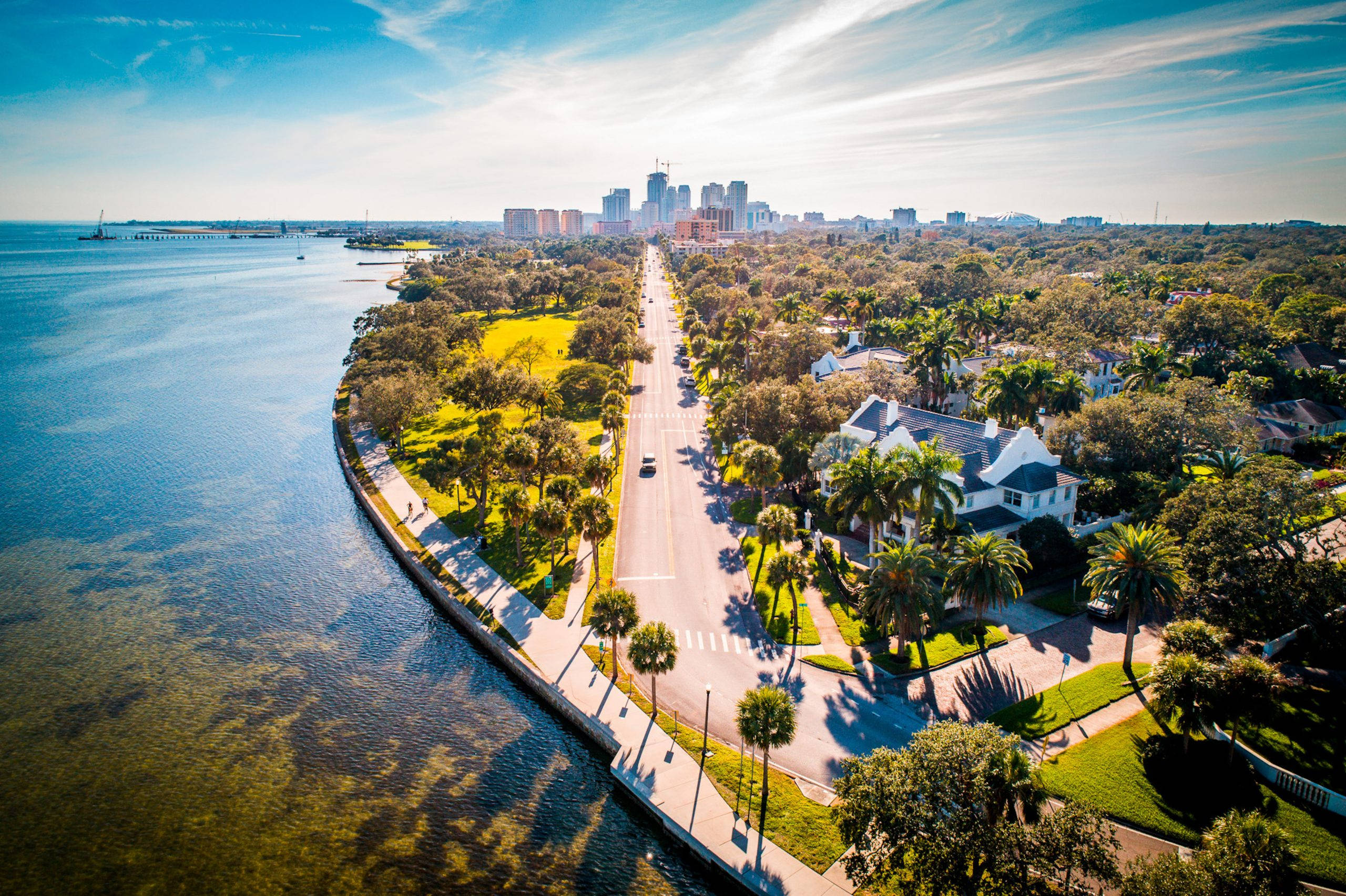 Road 桌布 壁紙 — 鳥瞰圖，一條沿海道路兩旁種滿棕櫚樹和房屋，通往遠處的城市天際線，天空湛藍明亮。