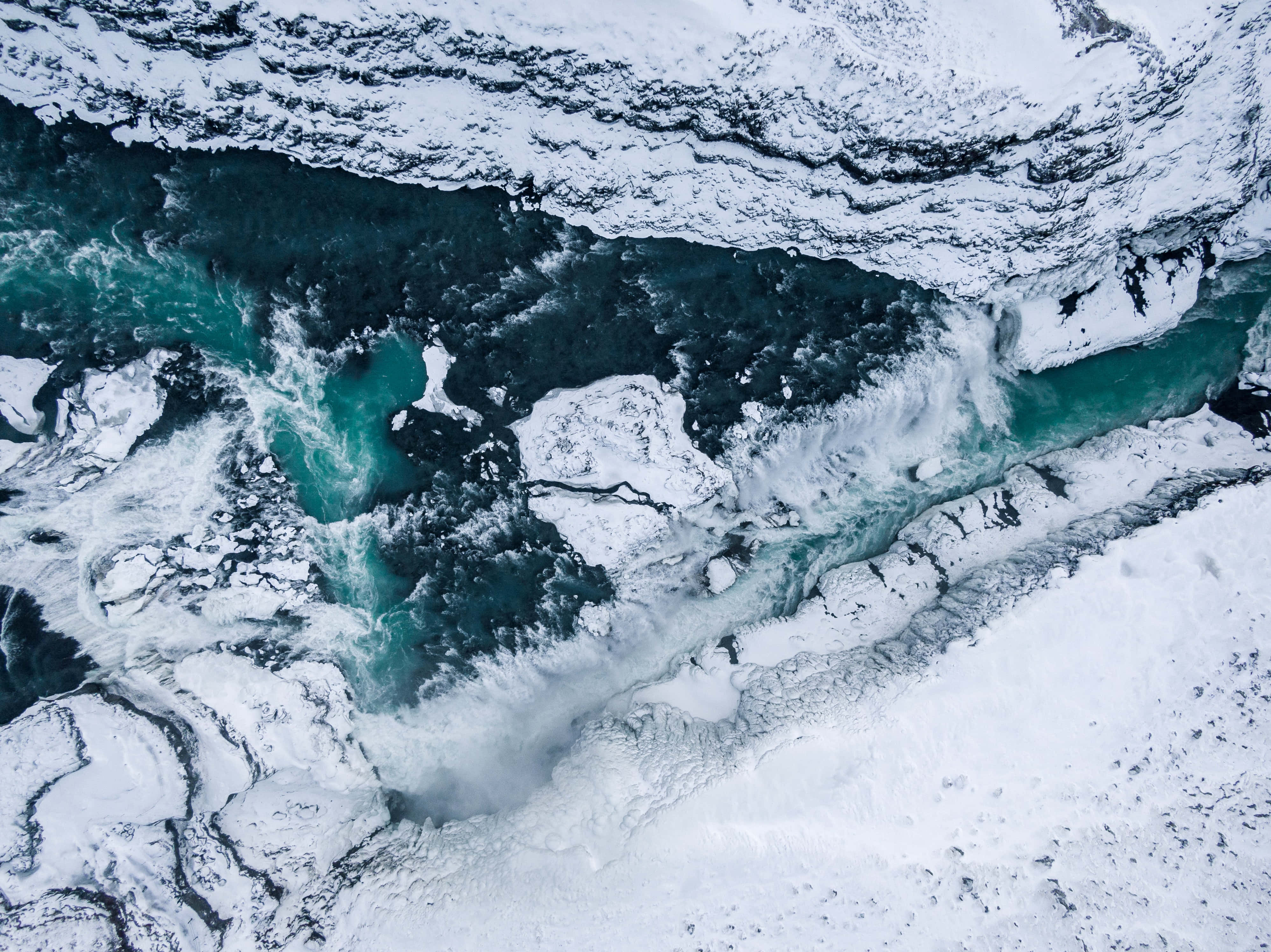 冰島 桌布 壁紙 — 鳥瞰圖：一條碧綠色的河流穿過白雪覆蓋的山脈，帶有漩渦狀的白色水流和冰質紋理。