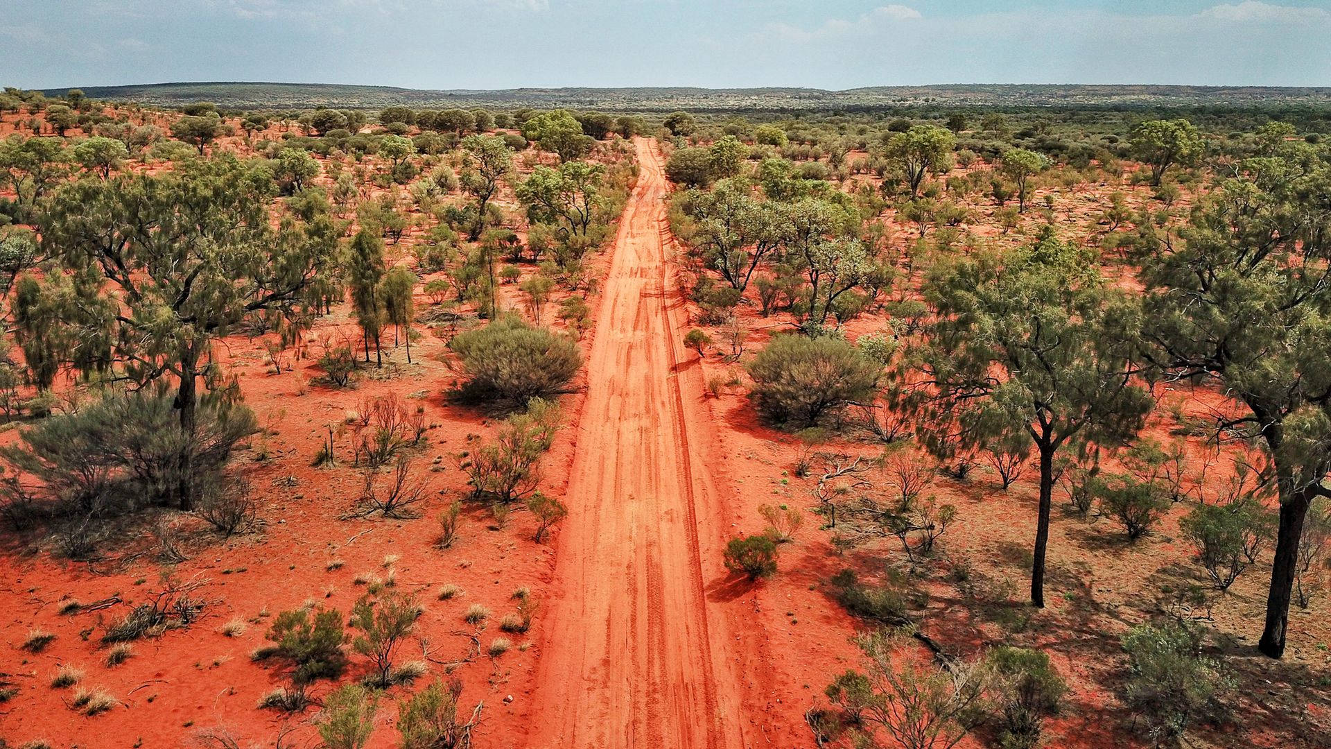 Outback 桌布 壁紙 — 鳥瞰圖：一條長長的紅色泥路穿過廣闊乾旱的內陸地區，散落著綠樹，天空清澈湛藍。