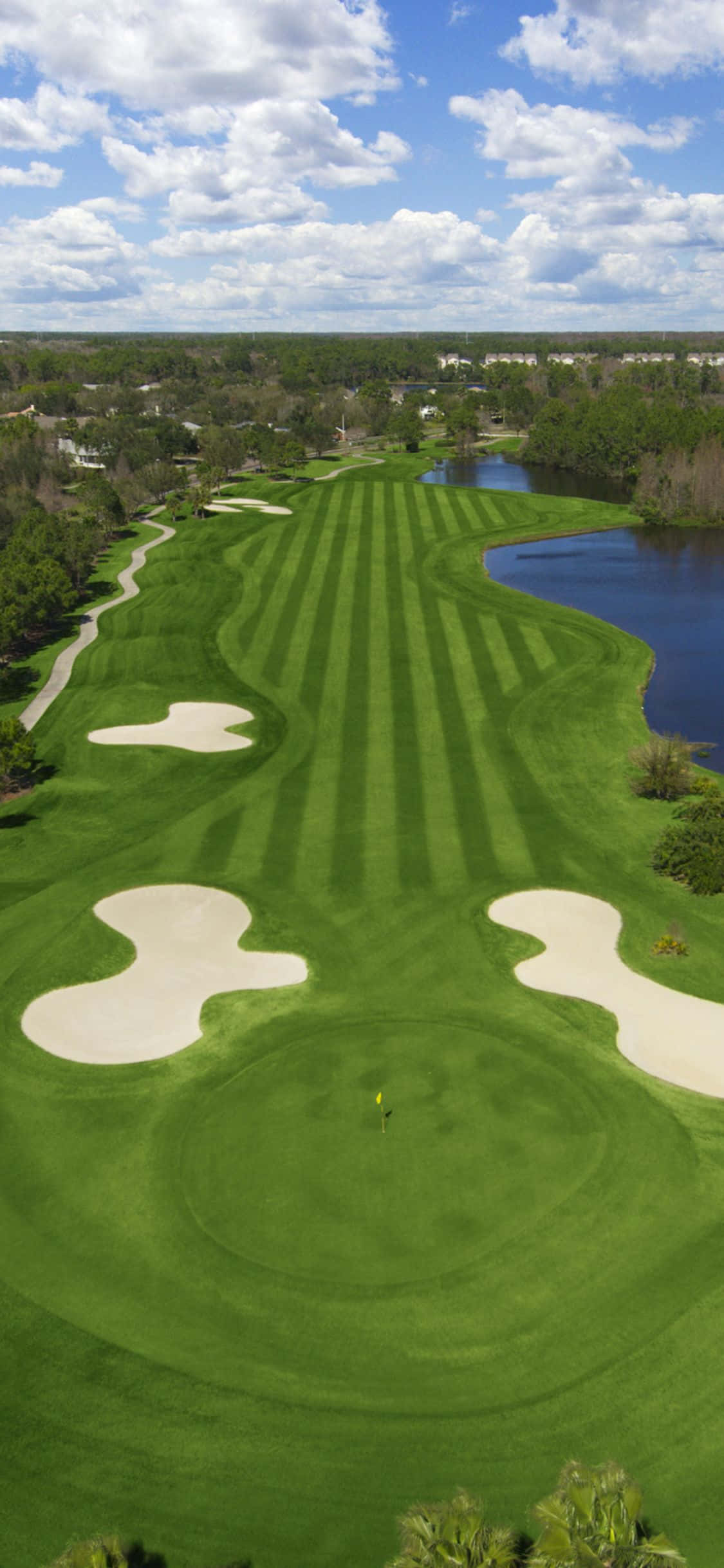 iPhone X Golf Course 桌布 壁紙 — 鳥瞰圖，一片翠綠的高爾夫球場，有條紋球道、沙坑和蜿蜒的水障礙，在明亮的藍天和白雲下。