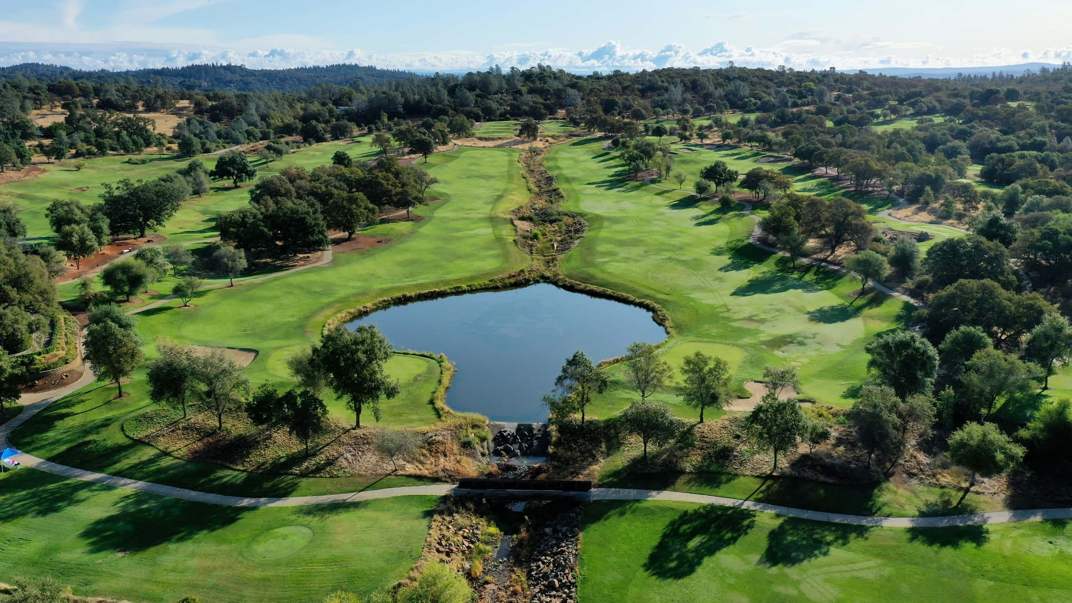 Golf Green 桌布 壁紙 — 鳥瞰圖：一片翠綠的高爾夫球場蜿蜒環繞著寧靜的池塘，四周樹木蔥蘢，遠山連綿，上方是湛藍的天空。