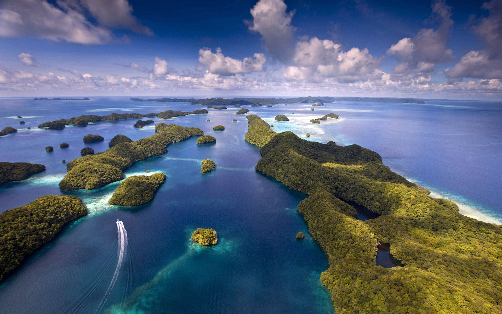 Palau 桌布 壁紙 — 鳥瞰圖：在明亮的天空下，翠綠的島嶼散落在充滿活力的碧綠和深藍色水域中，天空中有蓬鬆的雲朵。