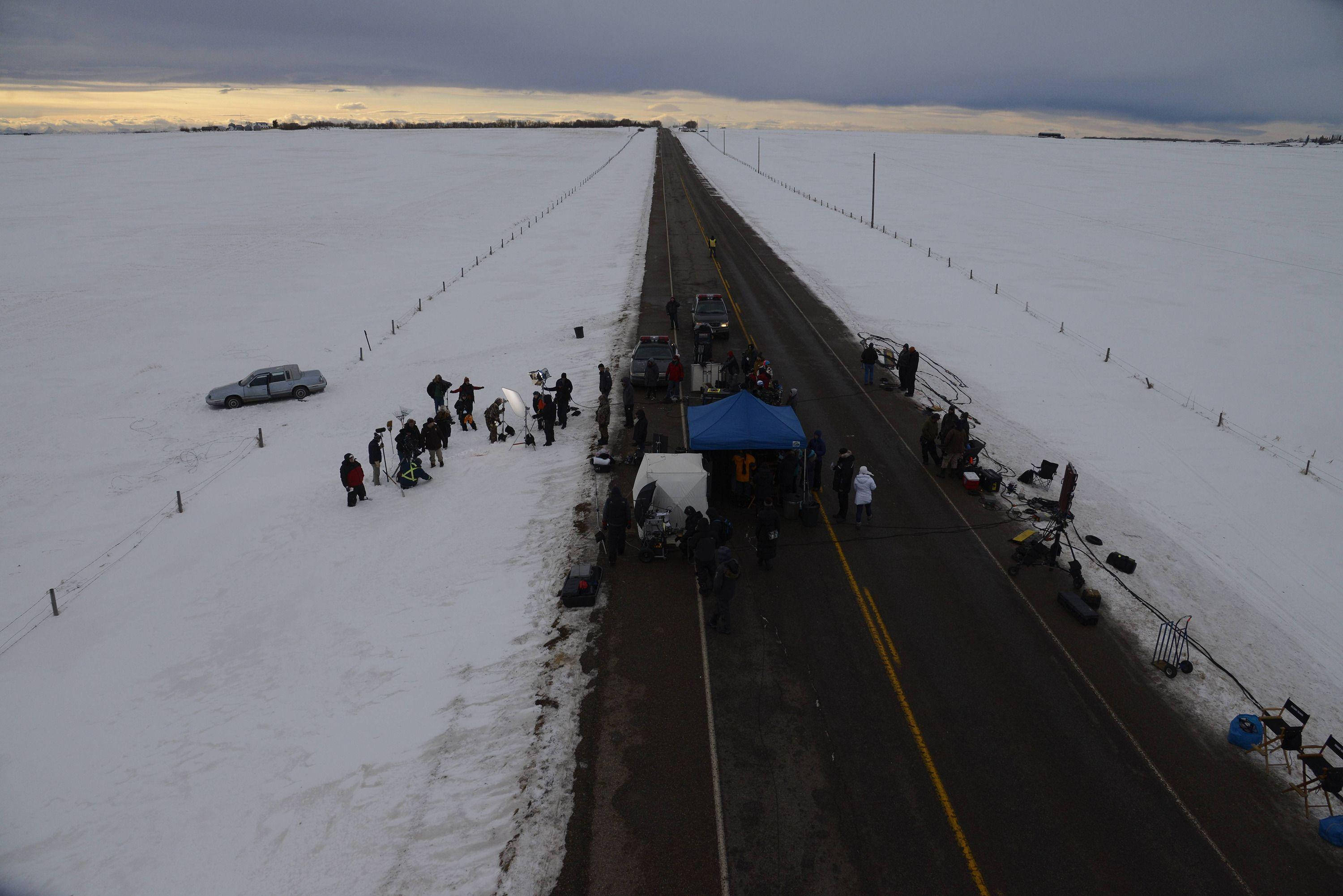 Road 桌布 壁紙 — 鳥瞰圖：在陰雲密布的黃昏天空下，一條積雪的道路延伸至廣闊的冰凍平原，路上聚集了人群和車輛。