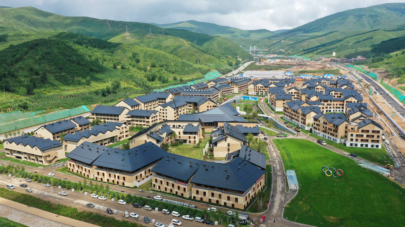 Winter Olympics 桌布 壁紙 — 鳥瞰圖：現代度假村建築，深色屋頂，坐落在鬱鬱蔥蔥的綠色山脈中，天空多雲。
