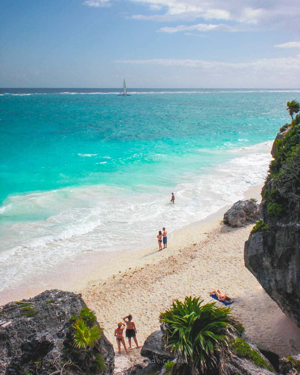 Mexican Beach 桌布 壁紙 — 鳥瞰圖：碧綠海洋與白色沙灘相接，周圍環繞著岩石峭壁，人們享受陽光，遠處有一艘帆船。