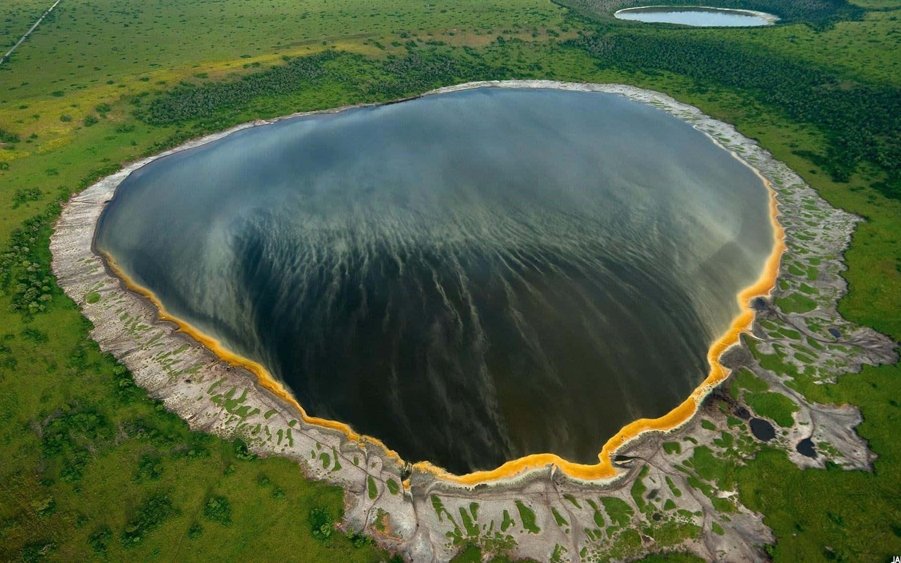 National Geographic Desktop 桌布 壁紙 — 鳥瞰圖顯示一個引人注目的圓形湖泊，湖水深色，邊緣有鮮黃色的礦物質，周圍是茂密的綠色地形。