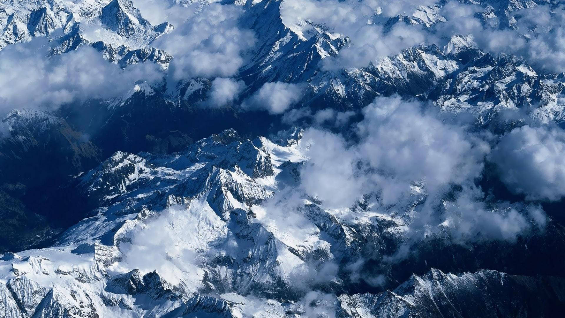 雪山 桌布 壁紙 — 鳥瞰圖顯示鋸齒狀的雪峰在明亮的日光下刺穿旋轉的白雲。