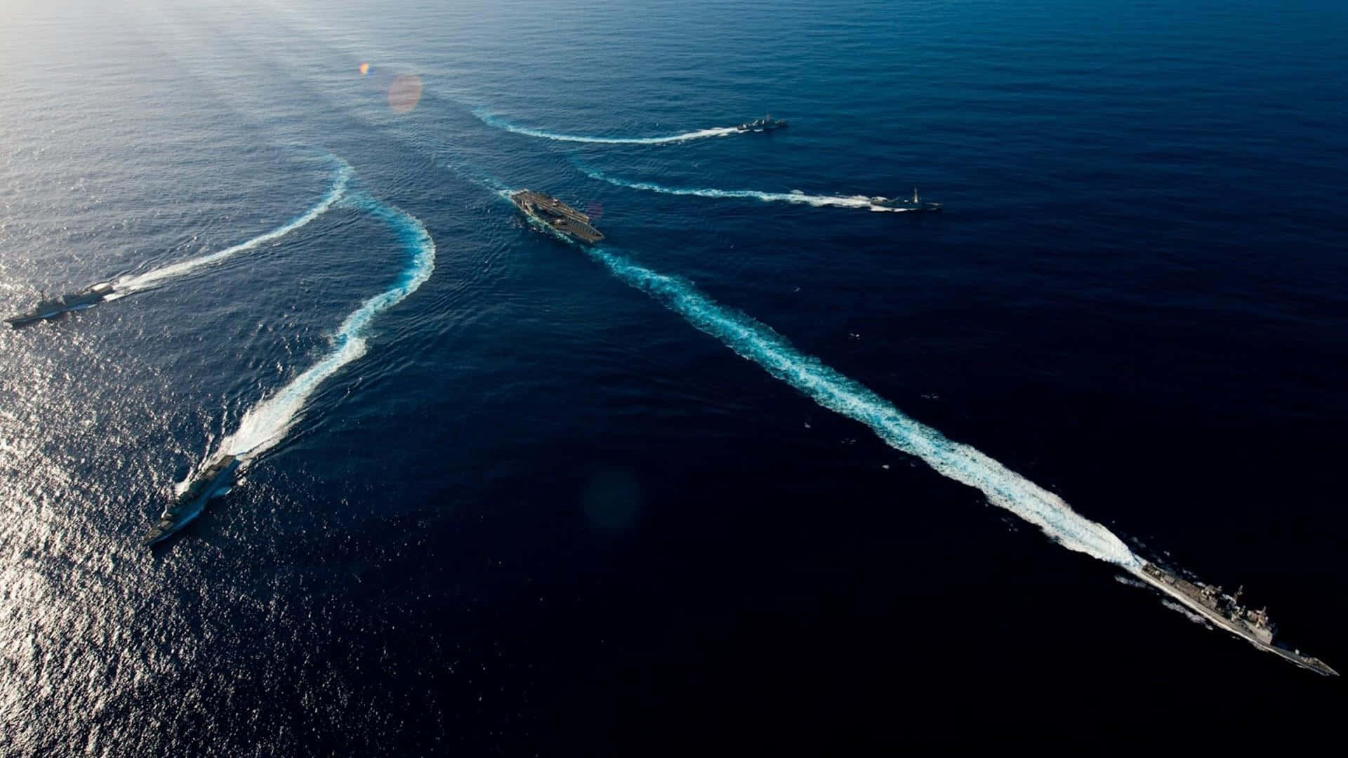 Navy 桌布 壁紙 — 鳥瞰海軍艦隊，包括航空母艦和驅逐艦，在深藍色海洋中破浪前行，在明亮的陽光下留下白色航跡。