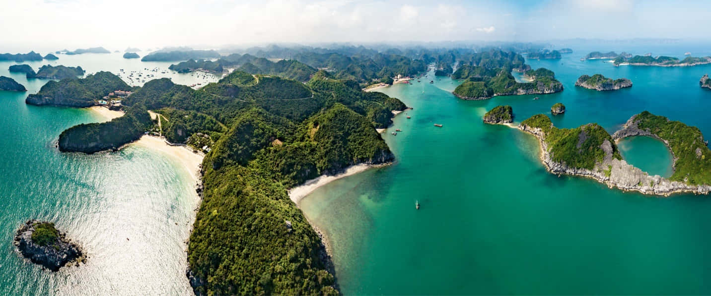 natural 桌布 壁紙 — 鳥瞰翡翠綠的海水環抱著翠綠的喀斯特島嶼，沙灣和散落的船隻在明亮的天空下。