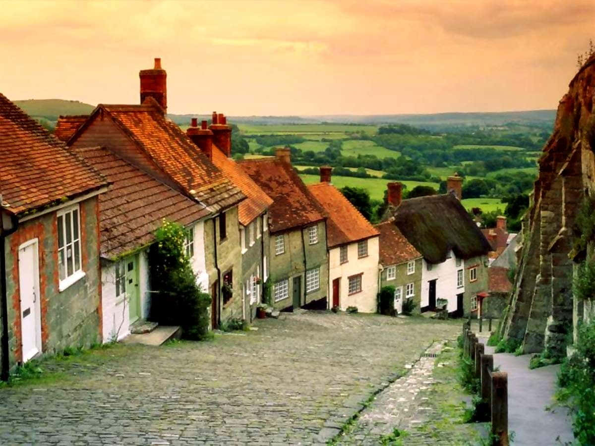England 桌布 壁紙 — 鵝卵石街道兩旁是歷史悠久的村舍和茅草屋頂，俯瞰著溫暖夕陽下連綿起伏的綠色山丘。