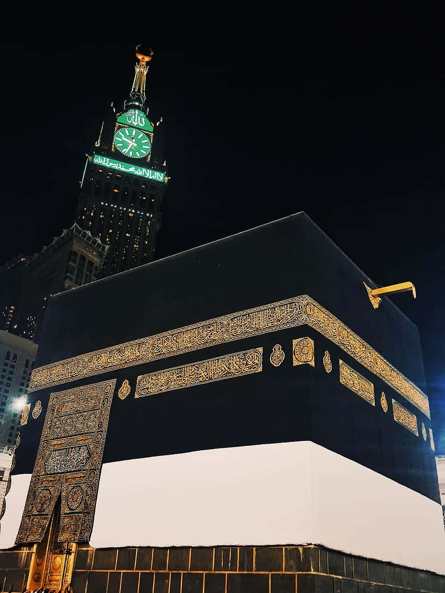 Makkah 桌布 壁紙 — 麥加的天房在夜晚被照亮，黑白布料上金色書法閃耀，背景中發光的鐘樓。