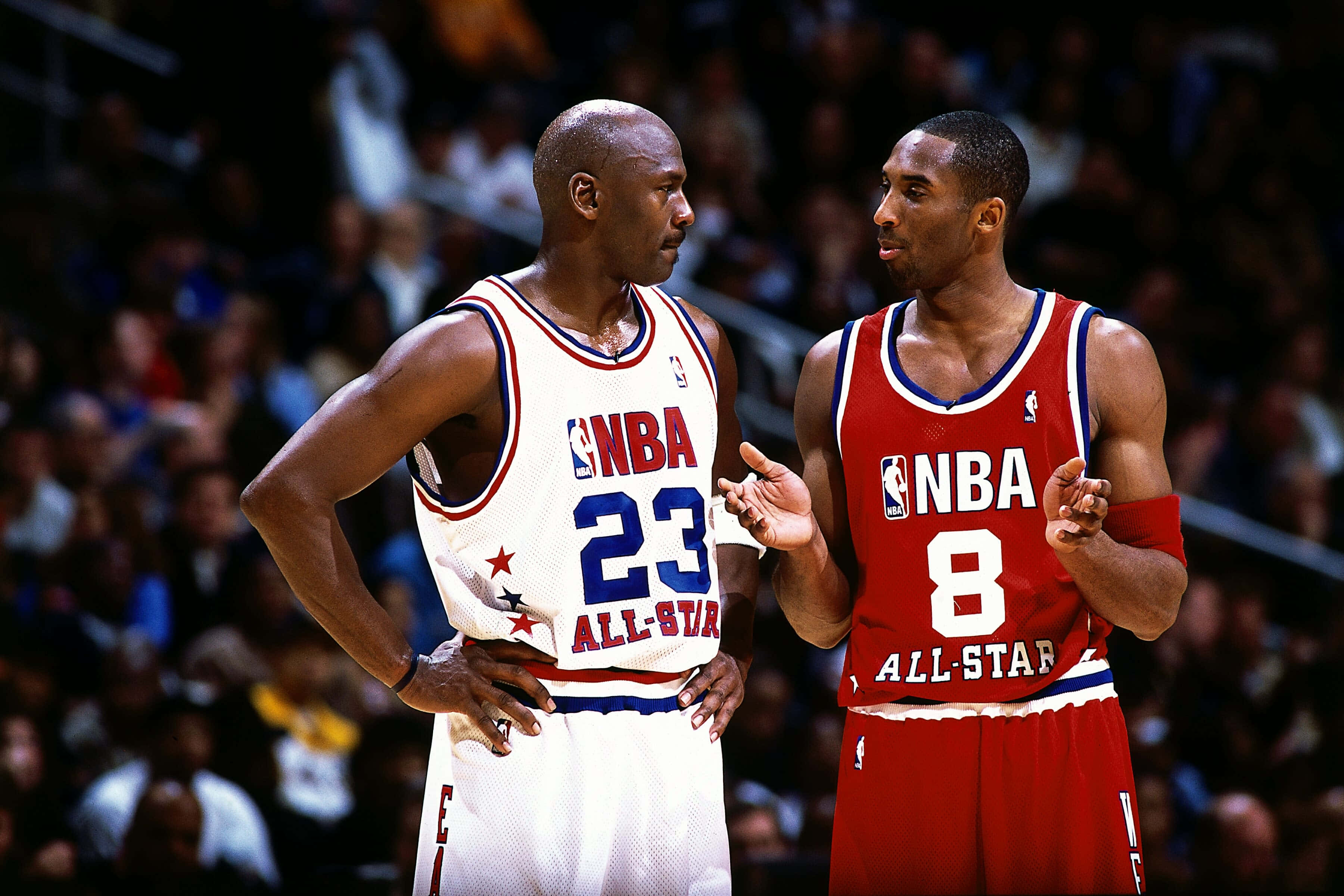 Kobe Bryant And Michael Jordan 桌布 壁紙 — 麥可喬丹和柯比布萊恩穿著NBA全明星球衣，在比賽中激烈交談，背景是模糊的人群。