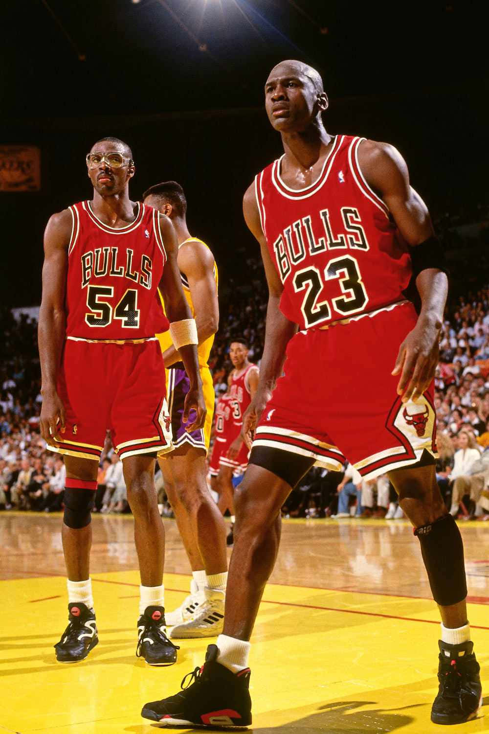 Jordan 桌布 壁紙 — 麥可喬丹身穿公牛隊23號球衣，在明亮的場館燈光下，與身穿54號球衣的隊友在籃球場上比賽時昂首挺立。