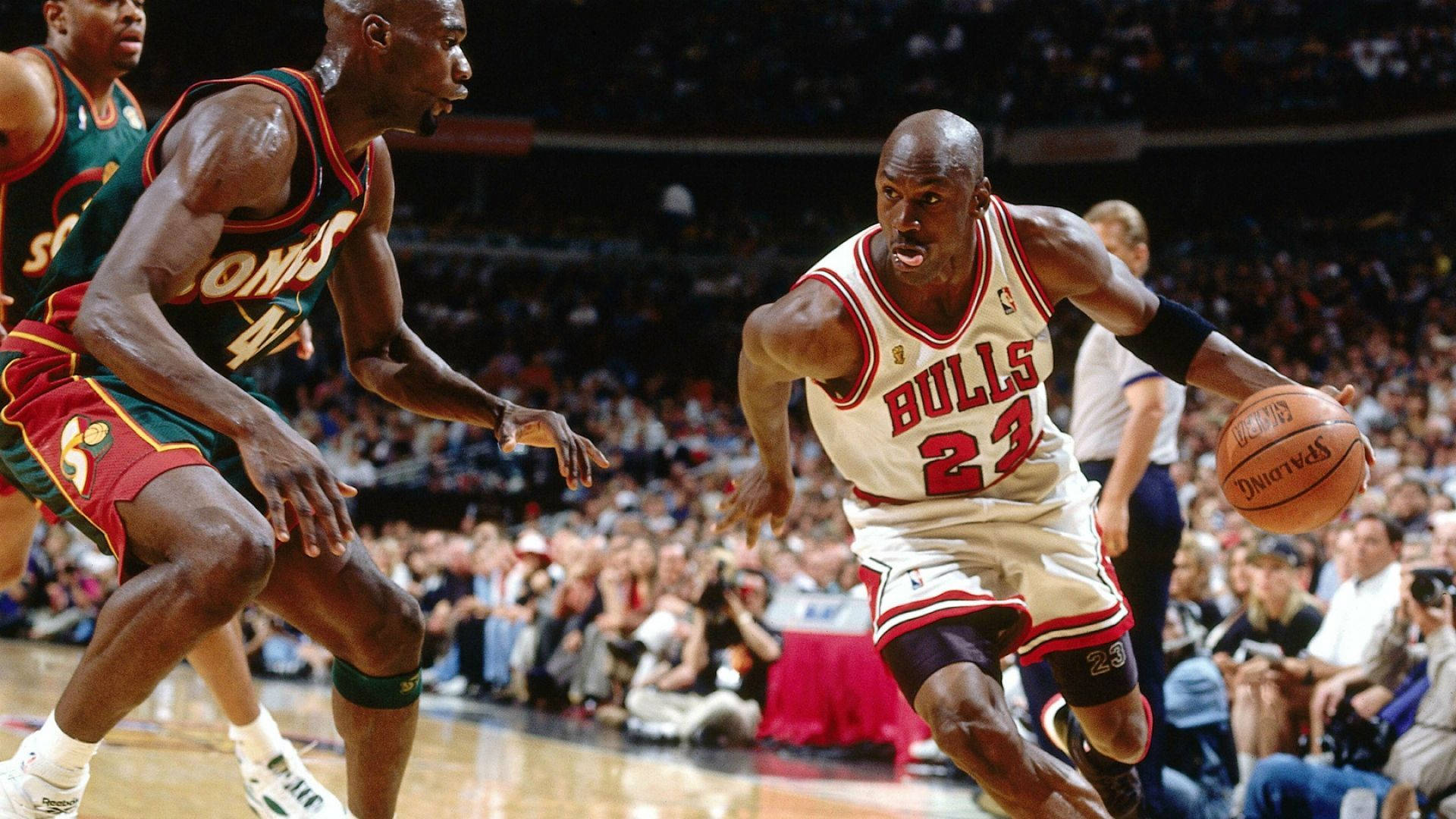 NBA 桌布 壁紙 — 麥可喬丹身穿白色公牛球衣，在場館燈光下的一場激烈籃球比賽中運球越過兩名防守球員。