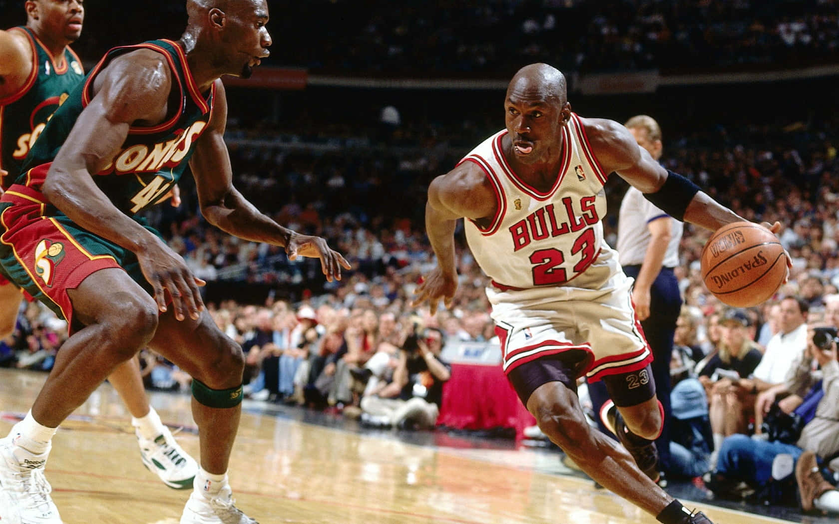 NBA 桌布 壁紙 — 麥可喬丹身穿白色公牛隊球衣，在球場燈光下於一場充滿活力的籃球比賽中運球越過防守球員。