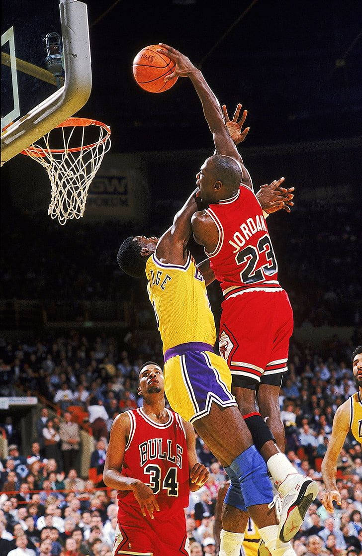 籃球 Jordan 桌布 壁紙 — 麥可喬丹身穿紅色公牛球衣，在擠滿觀眾的球場比賽中飛越湖人隊防守球員灌籃。