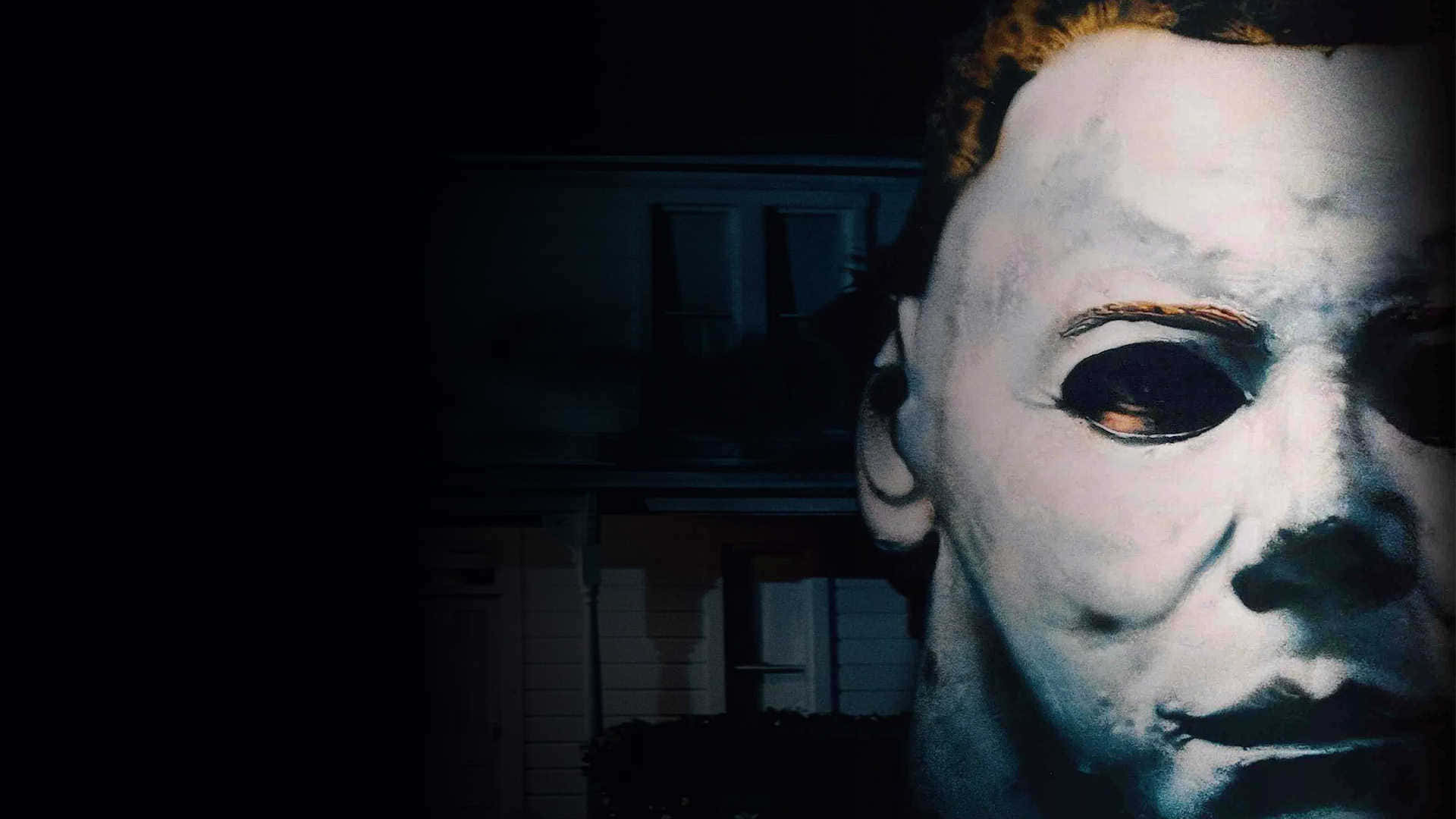 Mike Myers 桌布 壁紙 — 麥可邁爾斯標誌性白色面具的特寫，眼睛深邃，背景是夜間燈光昏暗的郊區房屋。