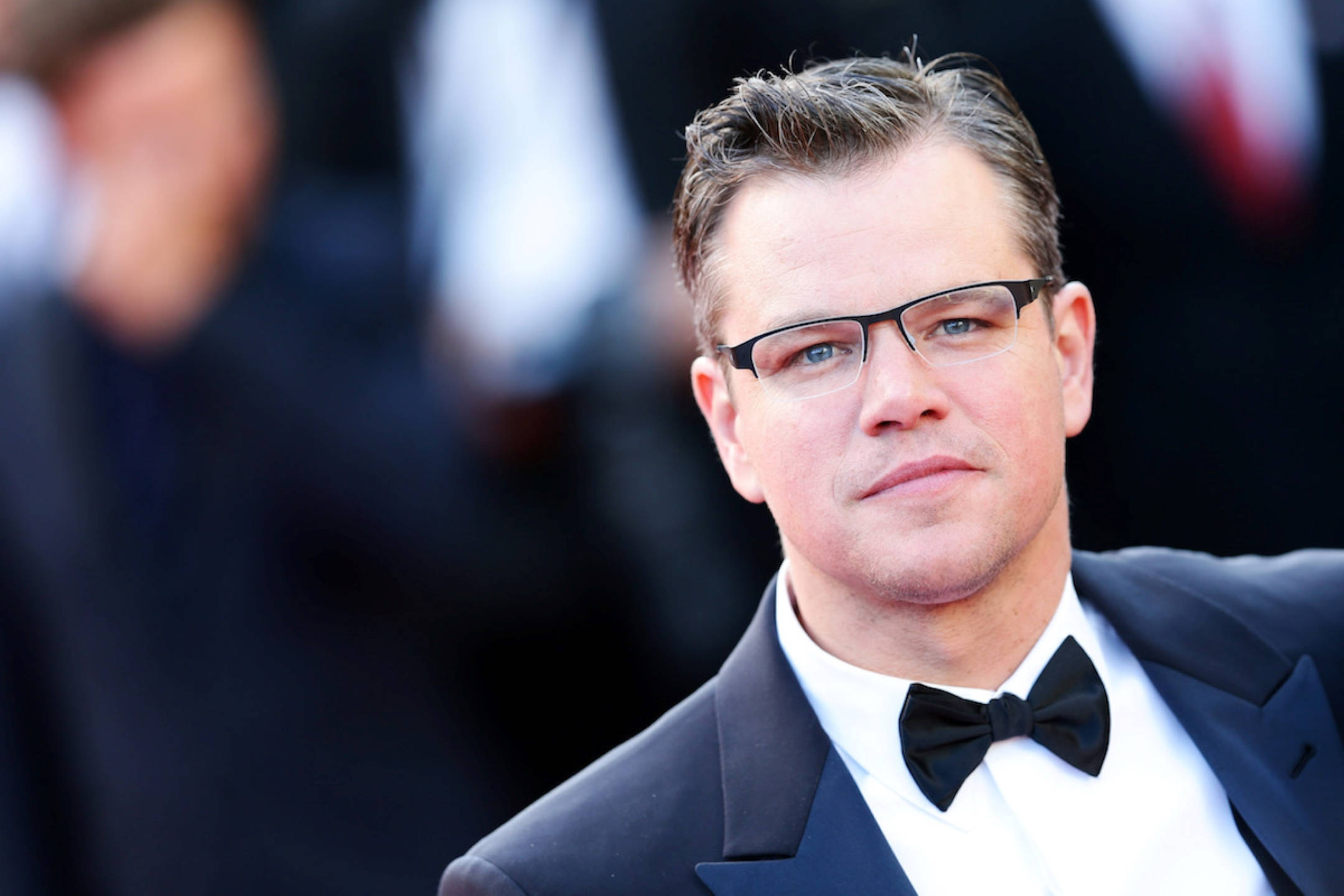 Matt Damon 桌布 壁紙 — 麥特戴蒙身穿俐落黑色禮服和領結，在明亮燈光下自信地站在紅地毯上擺姿勢。