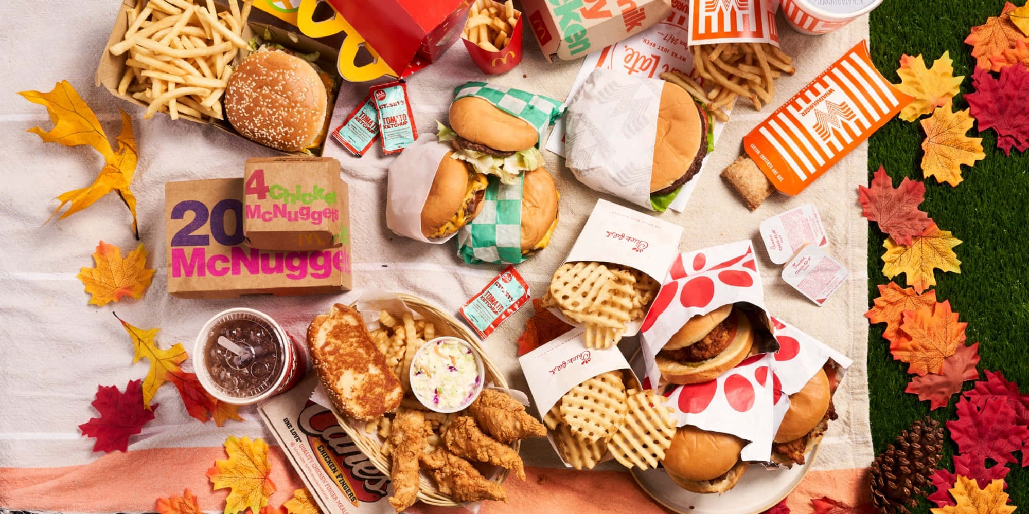 fast food 桌布 壁紙 — 麥當勞秋季主題食品（如麥克雞塊、漢堡、薯條和飲料）在毯子上與秋葉和松果一起擺放的俯視圖。