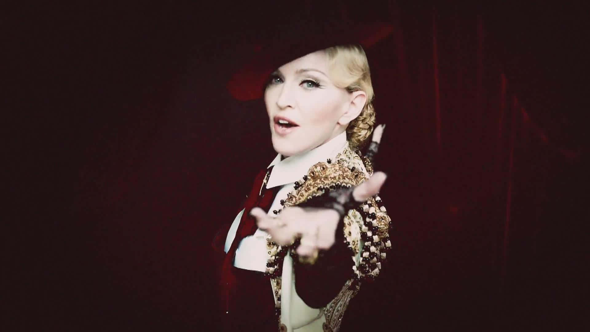 Madonna 桌布 壁紙 — 麥當娜穿著戲劇性的紅色和金色服裝，在深色天鵝絨背景和強烈的舞台燈光下向前指。