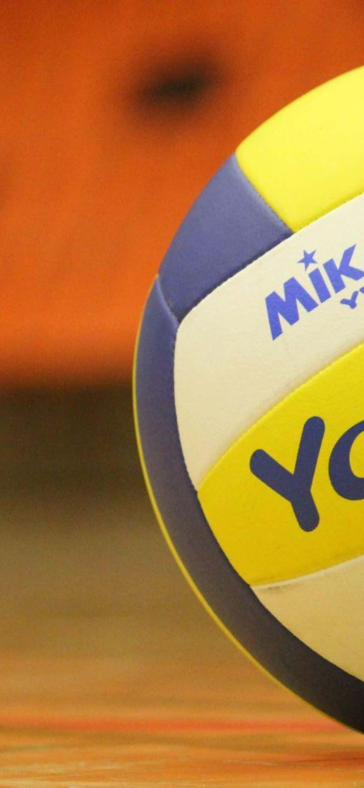 iPhone Xs Max Volleyball 桌布 壁紙 — 黃、藍、白三色 Mikasa 排球特寫，放置在木質球場上，背景溫暖模糊。