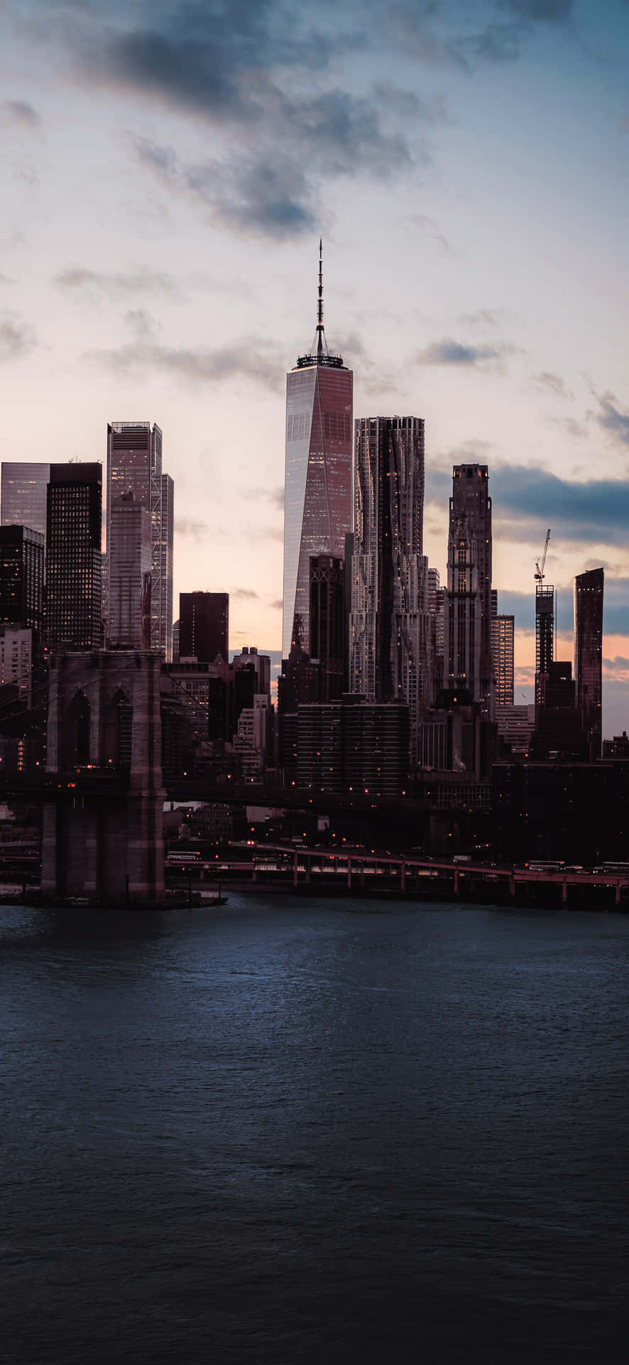 Skyscrapers next to a dark body of water 4K 壁紙 — 黃昏城市景觀，One World Trade Center 和布魯克林大橋在陰天天空下的水面上。