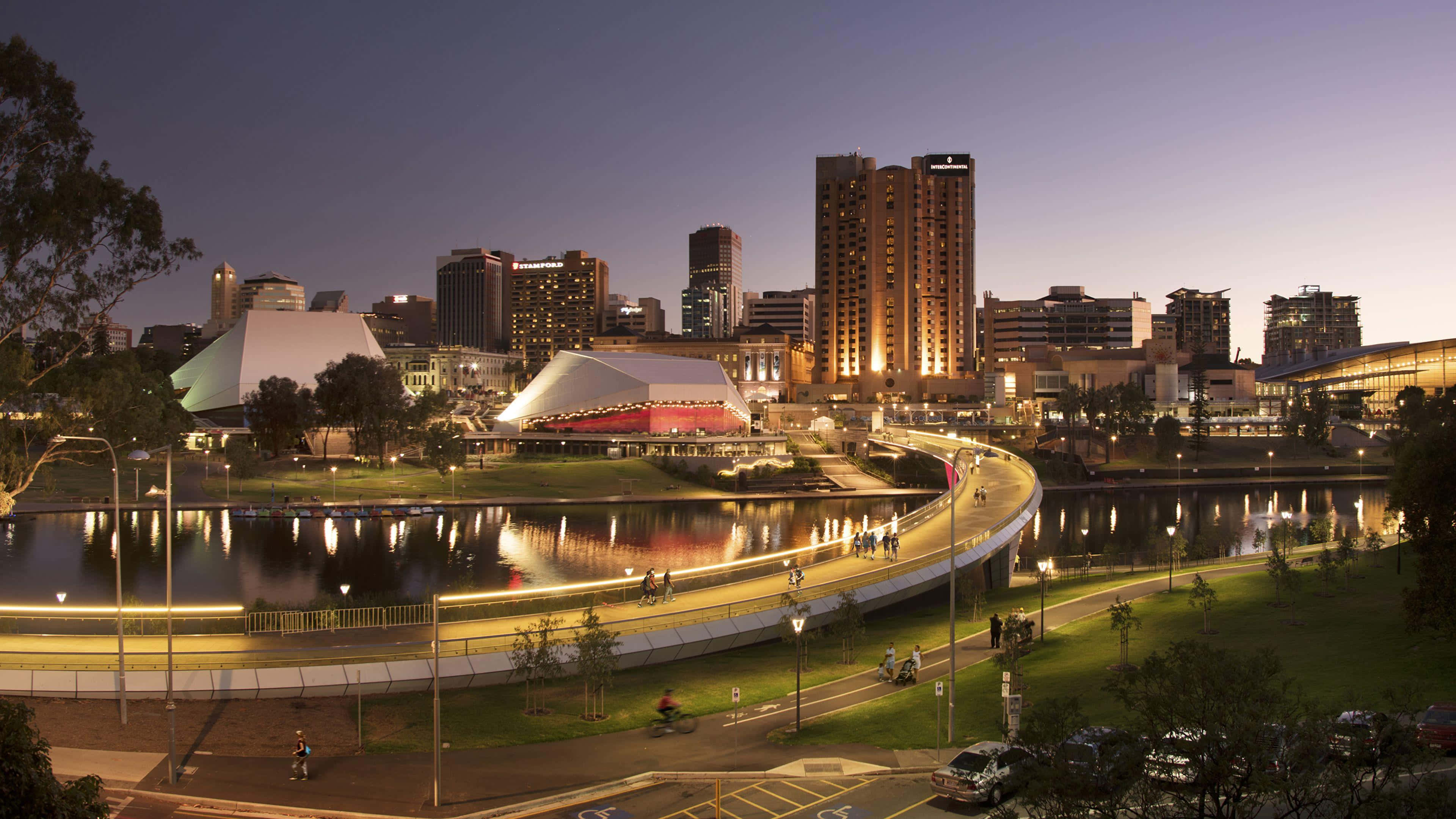 Adelaide 桌布 壁紙 — 黃昏城市景觀，有燈光璀璨的現代建築和彎曲的河濱步道，反射出溫暖的燈光。