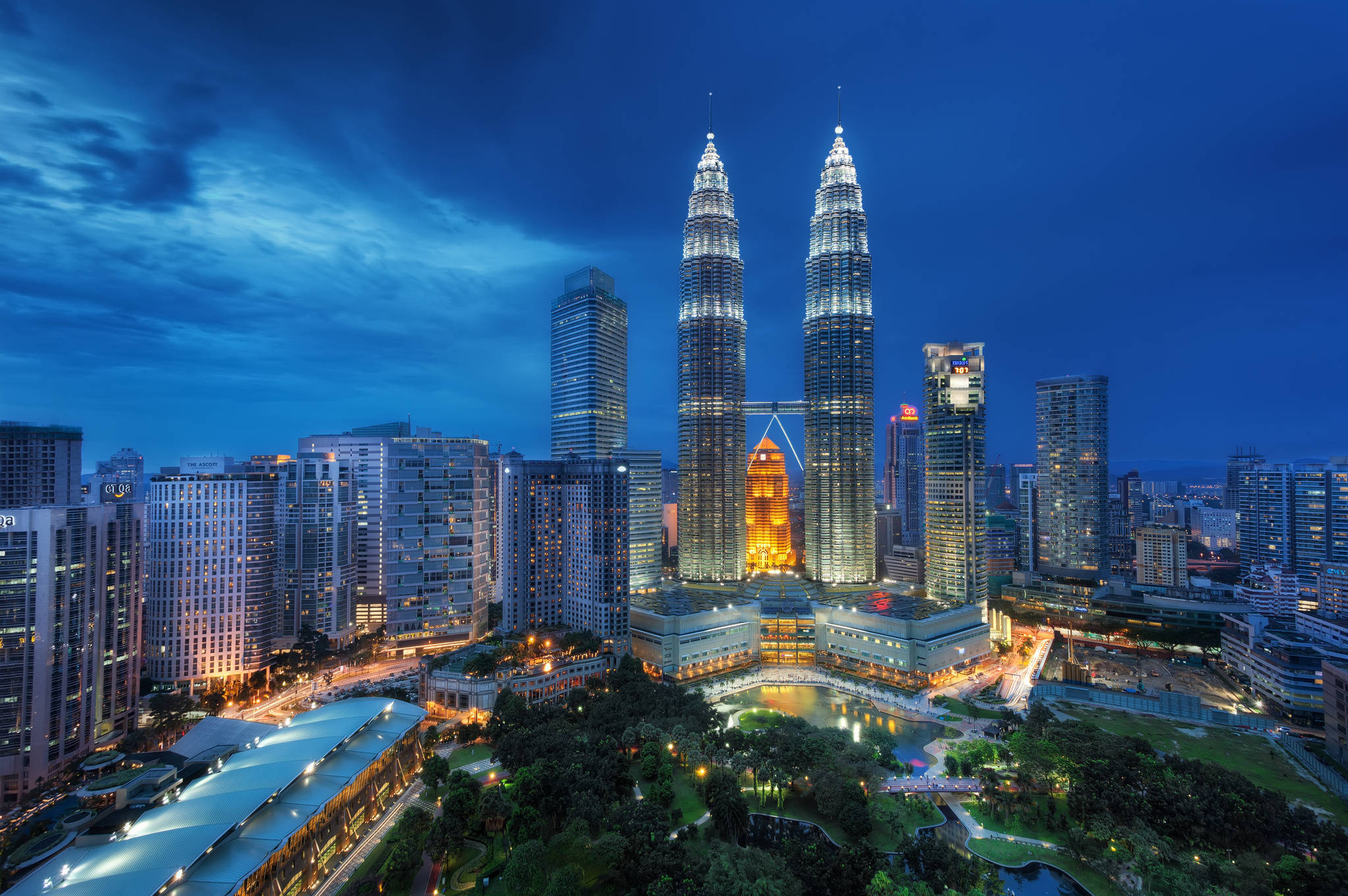 Malaysia 桌布 壁紙 — 黃昏城市景觀，標誌性的雙子星塔在深藍色天空中閃耀，周圍是高樓大廈和燈光點綴的綠化帶。