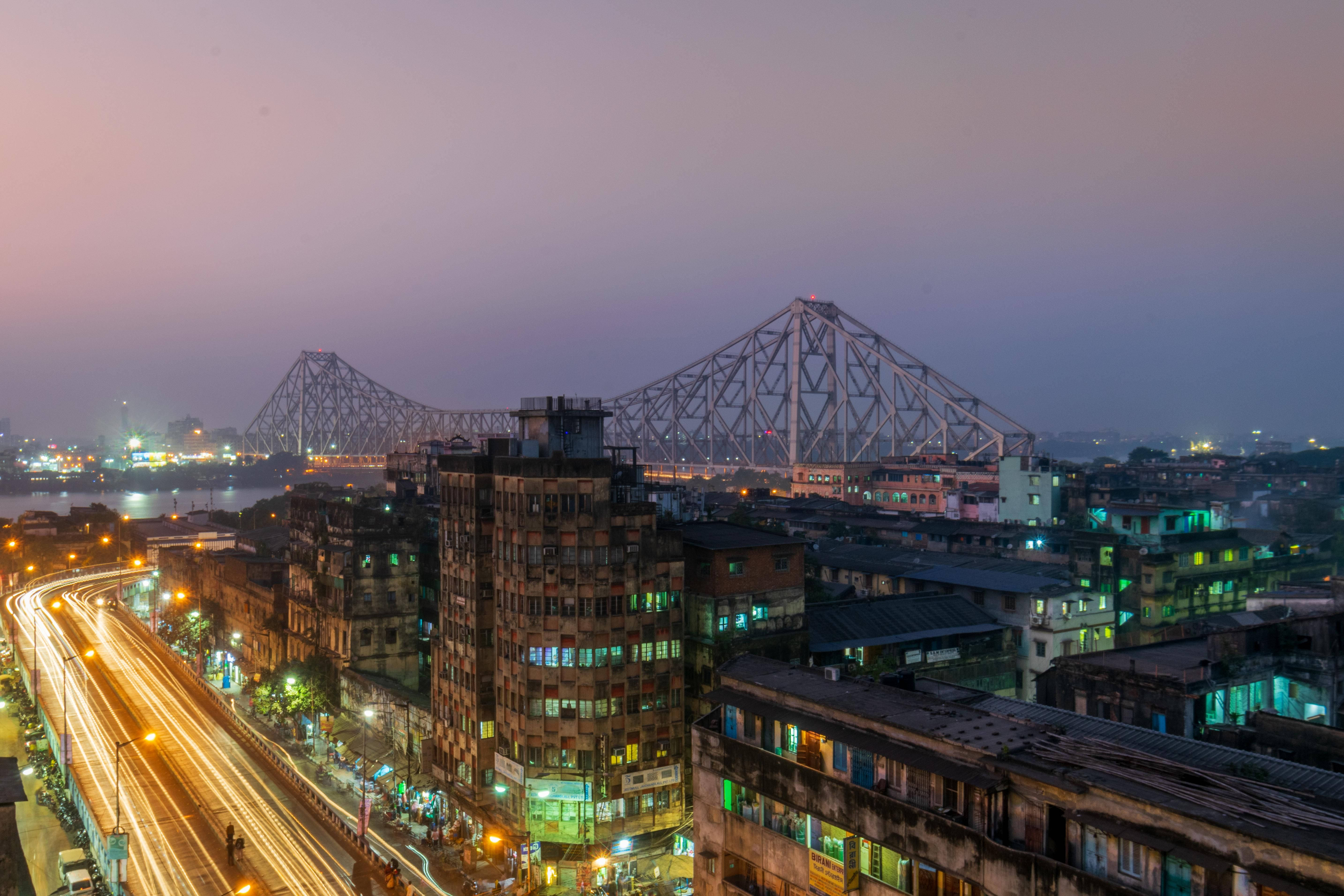 Kolkata 桌布 壁紙 — 黃昏城市景觀，水面上有一座發光的橋樑，帶有流動的交通燈光和明亮的建築物。