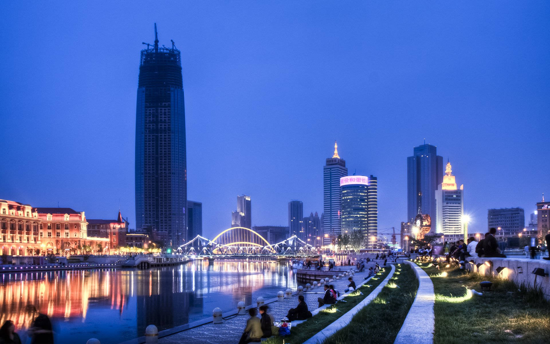 Tianjin 桌布 壁紙 — 黃昏城市景觀，燈火通明的摩天大樓和發光的橋樑倒映在平靜的水面上，人們沿著河邊小徑放鬆。