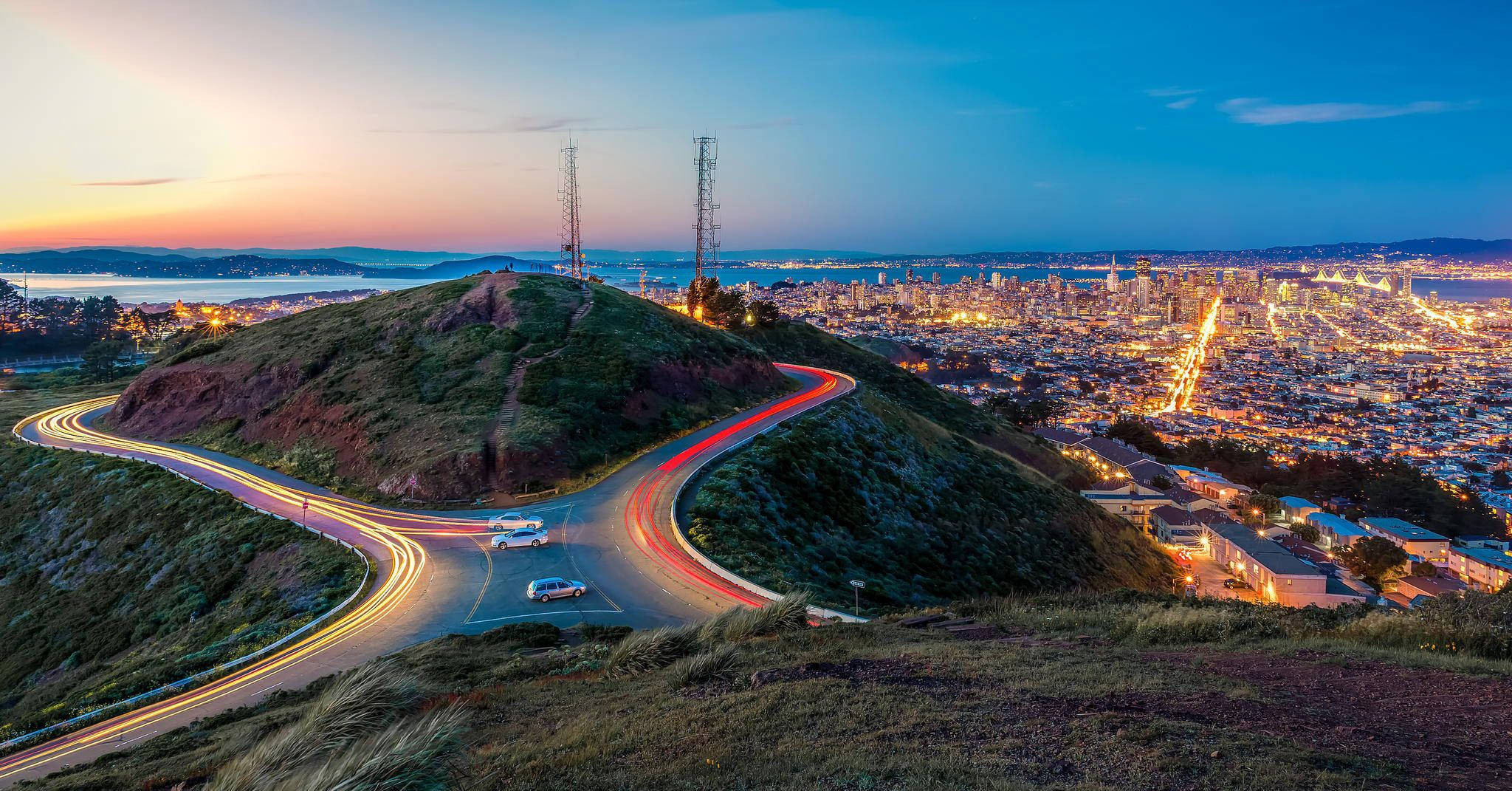 San Francisco 桌布 壁紙 — 黃昏城市景觀，發光的交通軌跡蜿蜒環繞著一個草坡，俯瞰廣闊的城市天際線。