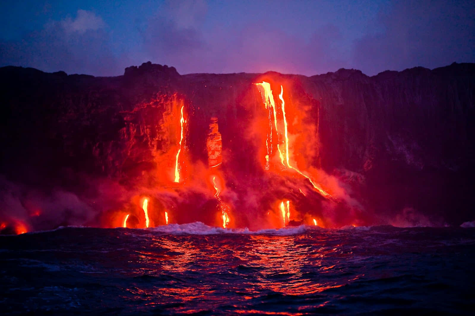 Volcano 桌布 壁紙 — 黃昏時，熔岩從懸崖流入海洋，在黑暗的海浪上投射出熾熱的紅光。