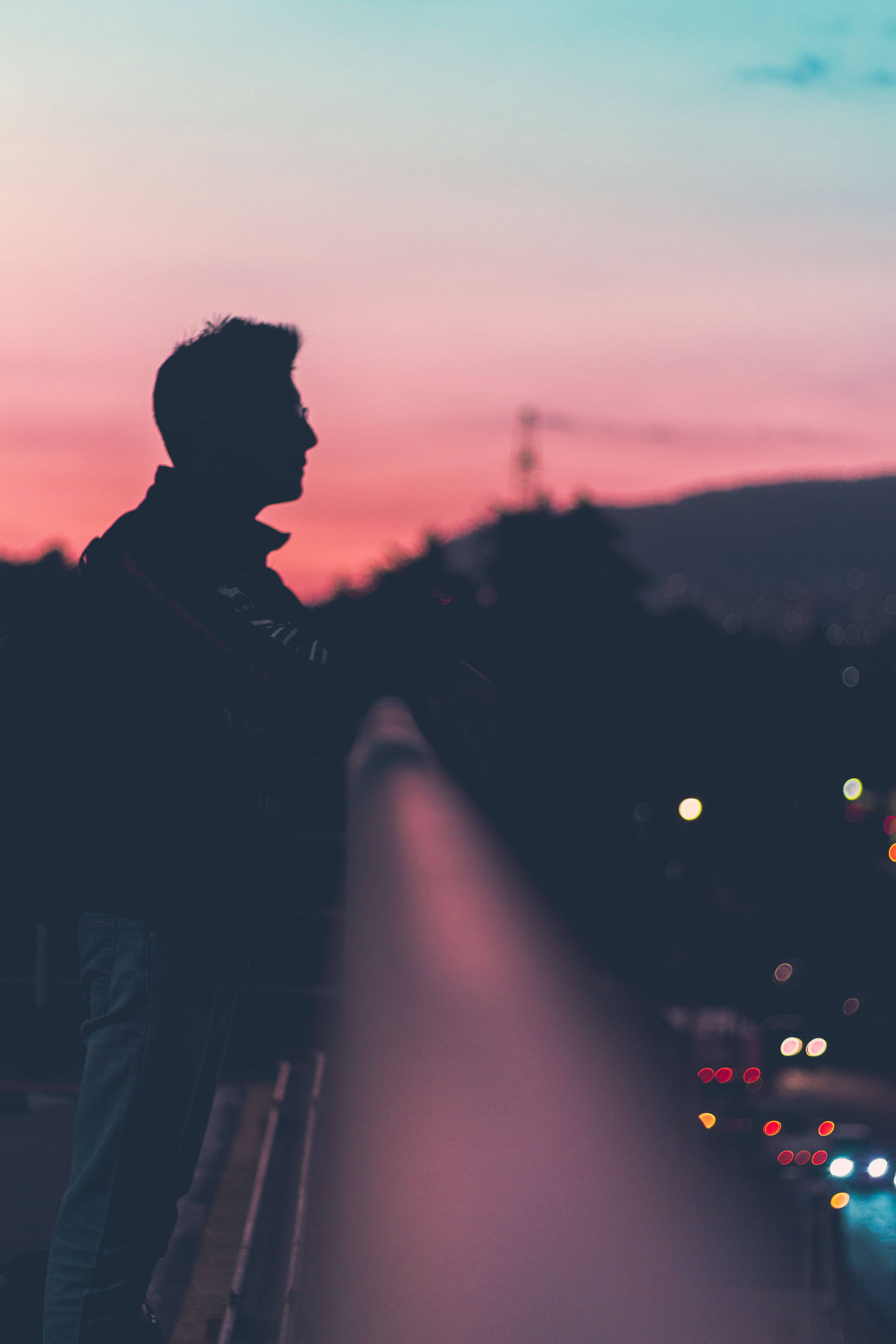 Silhouette 桌布 壁紙 — 黃昏時分，一個人站在橋上的剪影，背景是模糊的城市燈光和粉橙色天空。