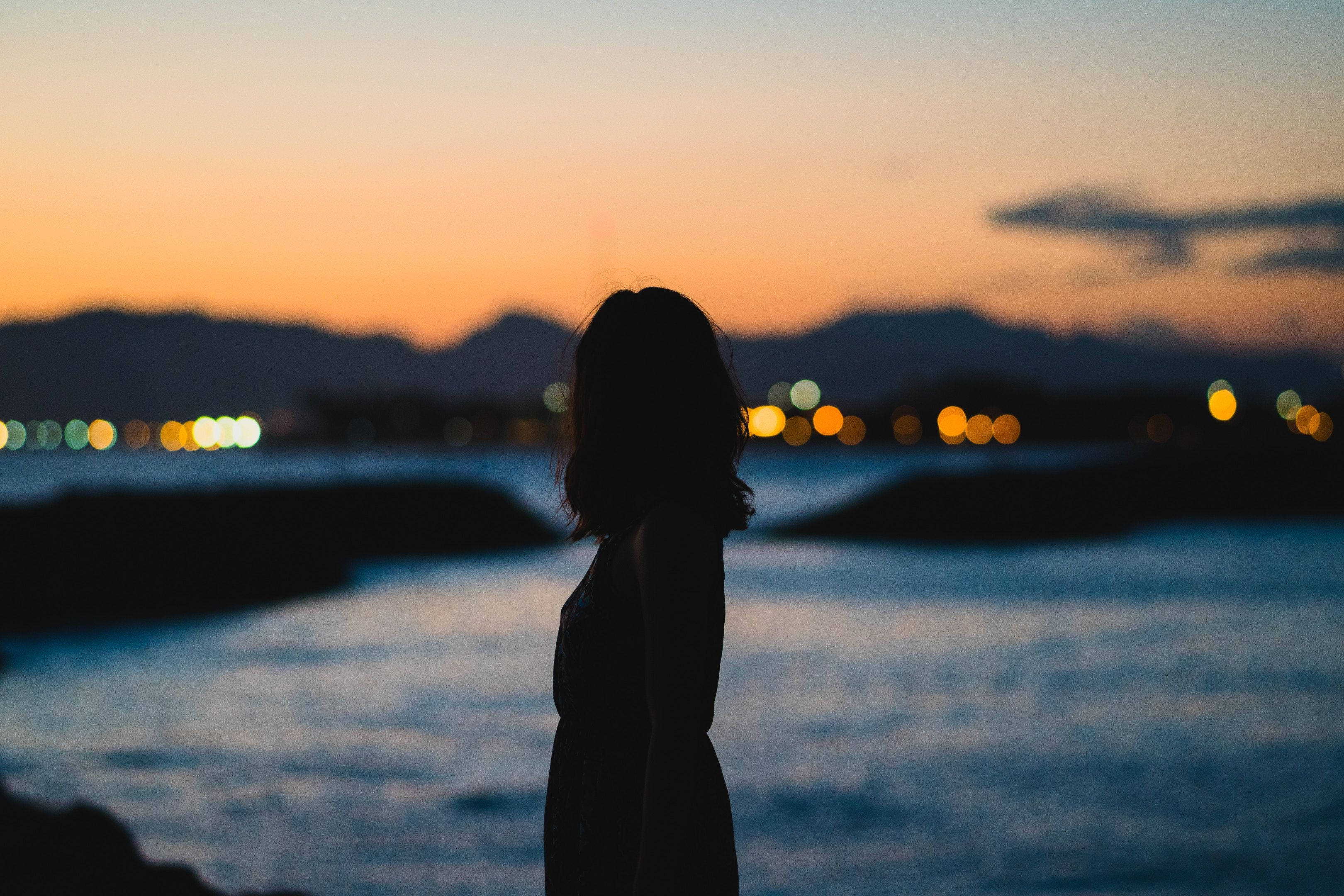 Woman Silhouette 桌布 壁紙 — 黃昏時分，一個人站在水邊的剪影，背景中遠處的城市燈光柔和地閃爍。
