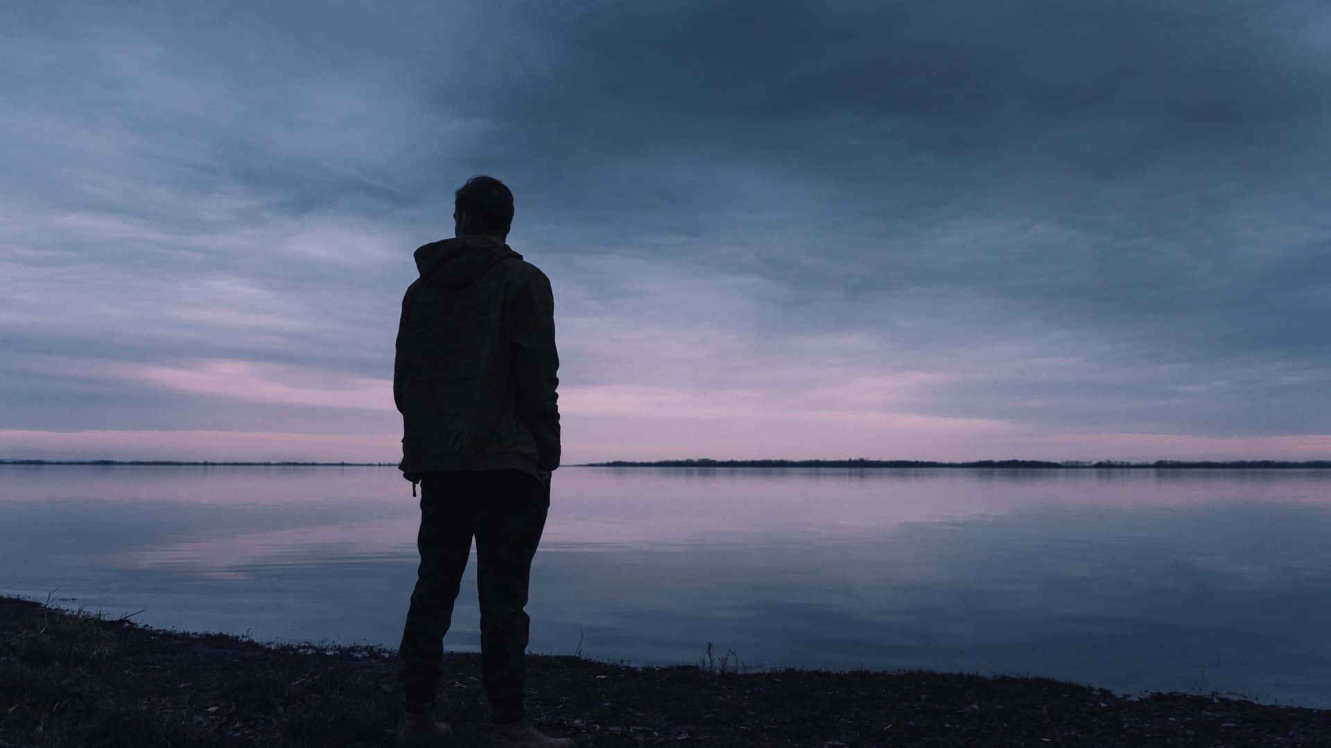 Lonely 桌布 壁紙 — 黃昏時分，一個孤獨的身影在湖岸上形成剪影，凝視著平靜的水面，天空呈現憂鬱的紫色和灰色。