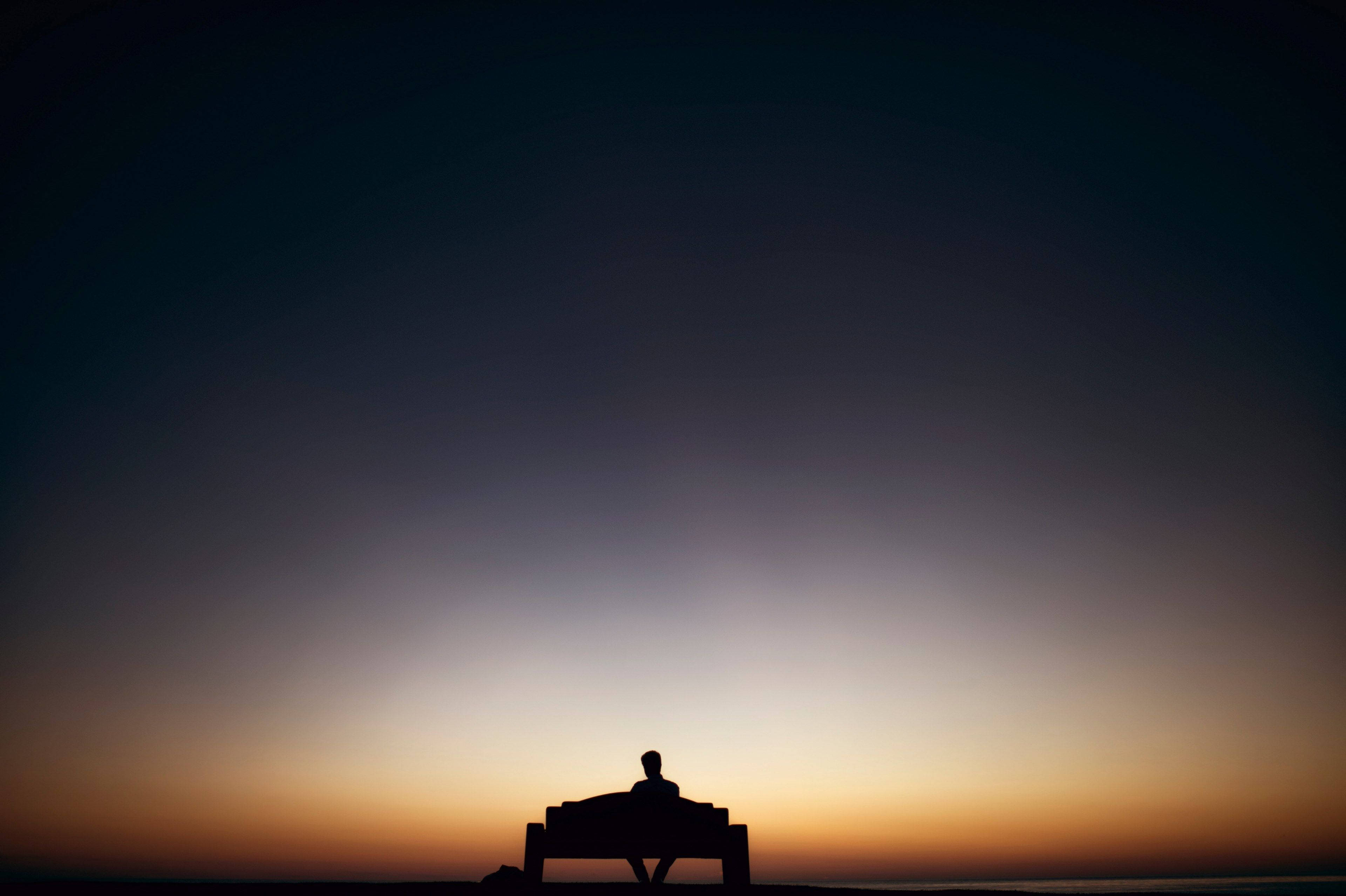 Silhouette 桌布 壁紙 — 黃昏時分，一個孤獨的身影坐在長椅上，背景是從深藍到暖橙的漸變天空。