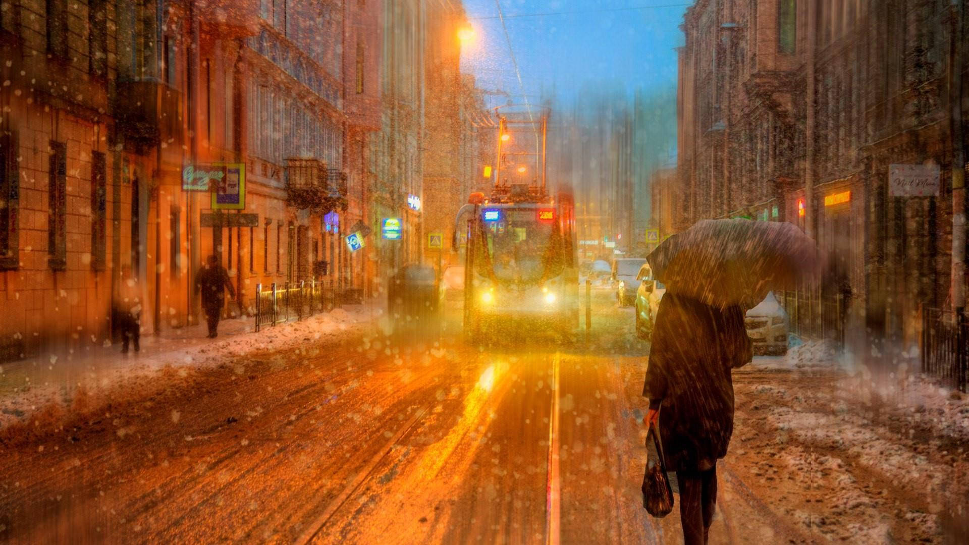 Saint Petersburg 桌布 壁紙 — 黃昏時分，一個孤獨的身影撐著傘走在積雪的都市街道上，電車的頭燈穿透飄落的雪花和街燈溫暖的光芒。
