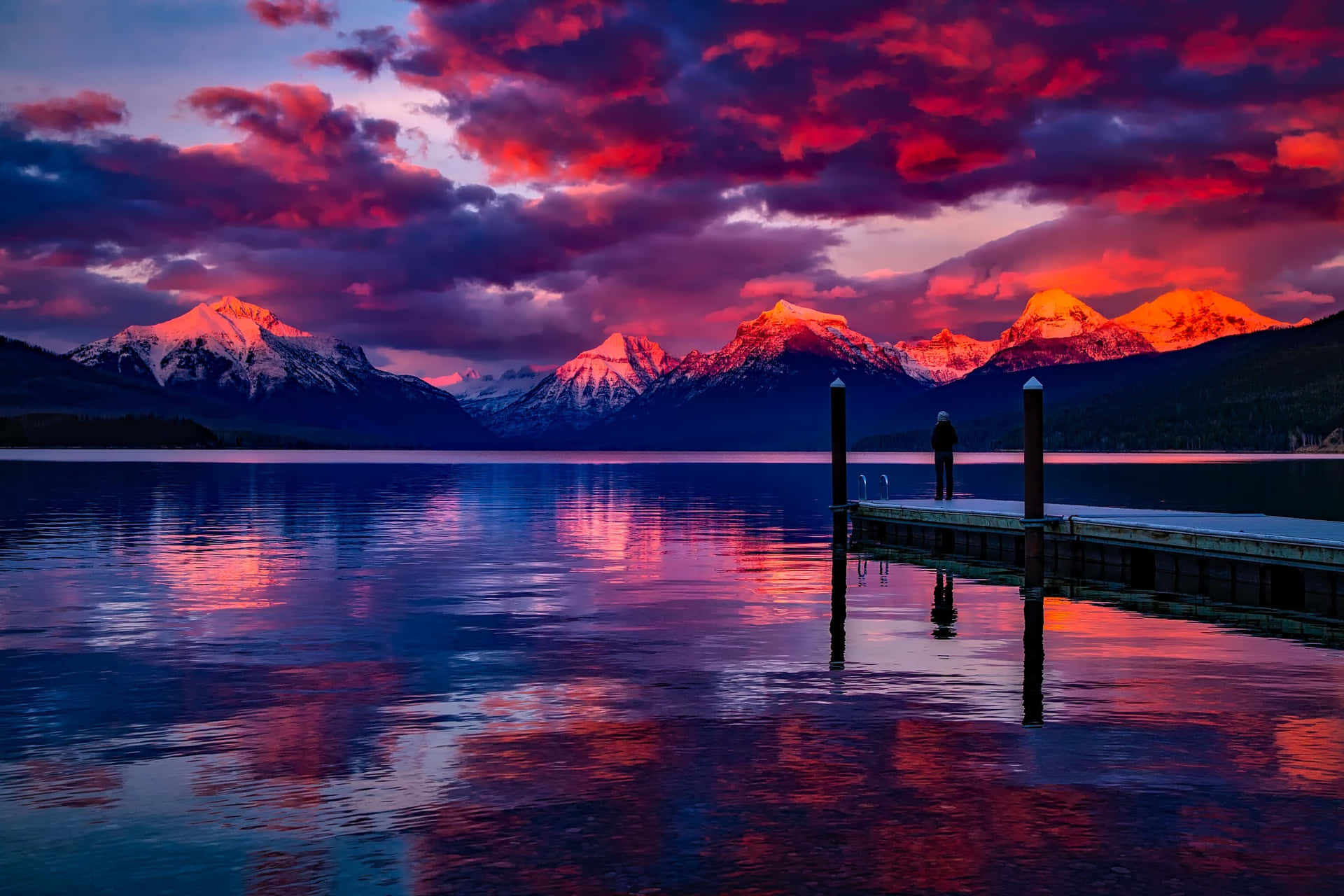 漂亮的動圖 桌布 壁紙 — 黃昏時分，一個孤獨的身影站在木製碼頭上，火紅和紫色的雲彩倒映在平靜的湖面上，四周環繞著積雪的山峰。