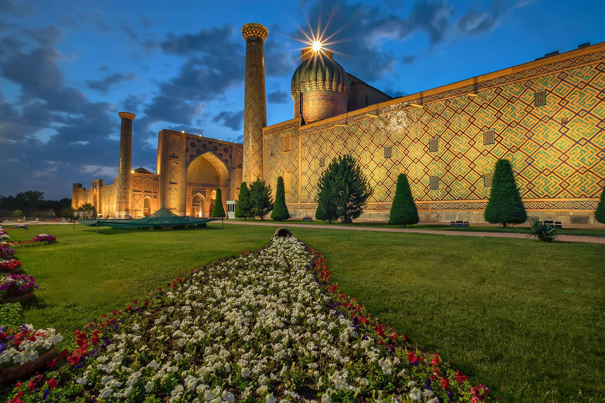 Uzbekistan 桌布 壁紙 — 黃昏時分，一個宏偉的伊斯蘭建築群在星空下亮起燈光，周圍是色彩繽紛的花壇和修剪整齊的花園。