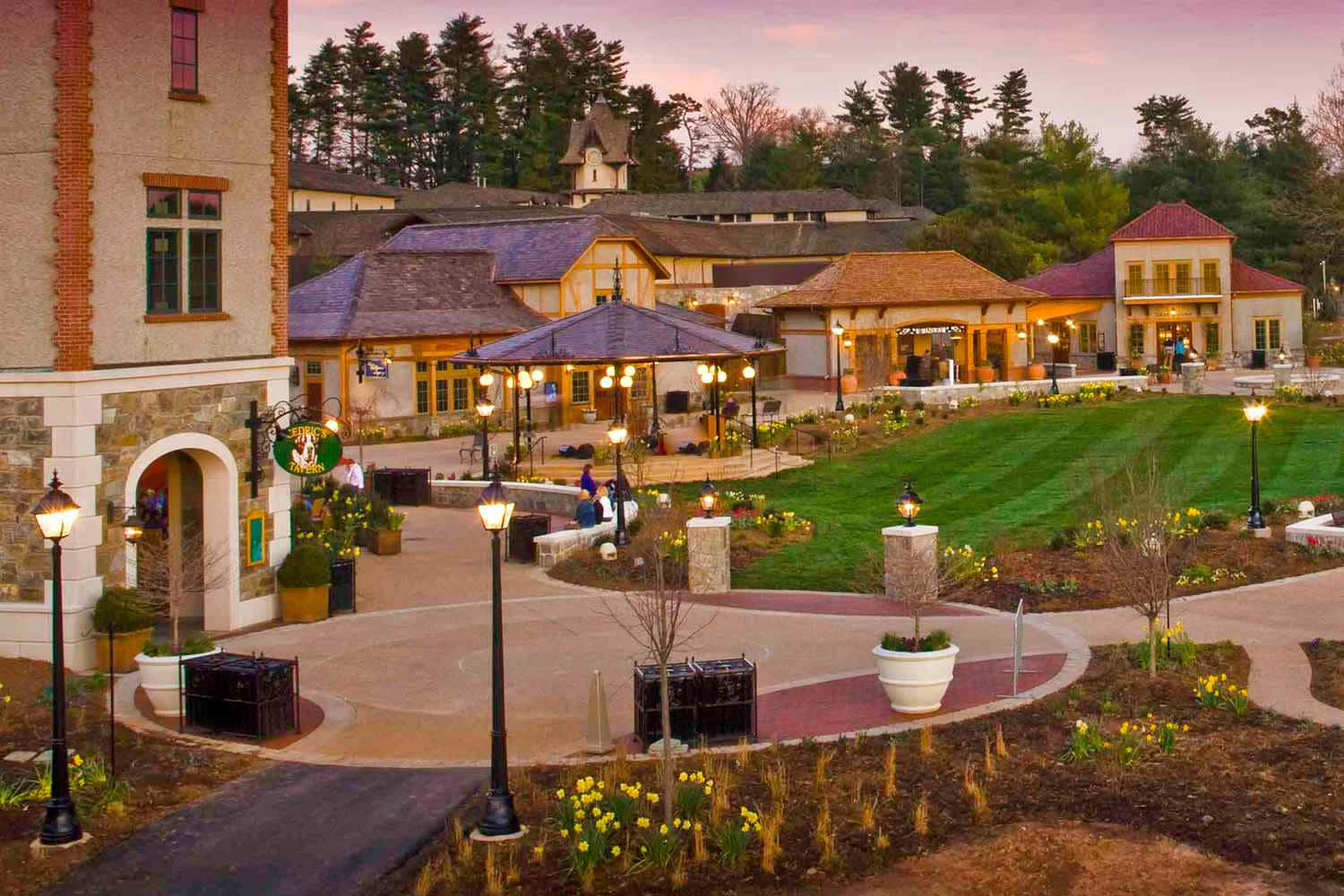 Biltmore Estate 桌布 壁紙 — 黃昏時分，一個宏偉的度假村綜合體，窗戶和露台散發出溫暖的燈光，周圍環繞著修剪整齊的草坪和盛開的黃色花朵。