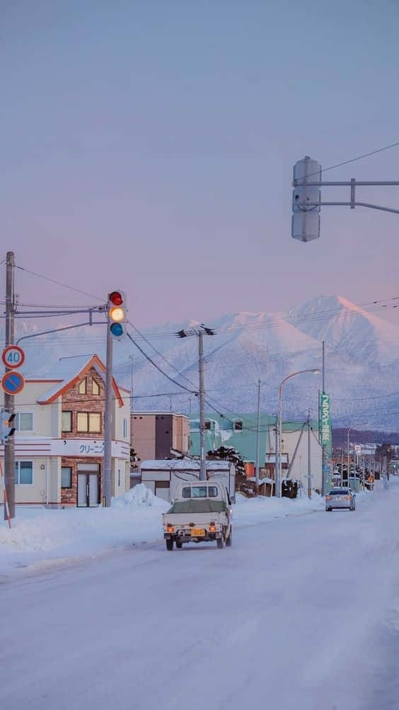 Snow iPhone 桌布 壁紙 — 黃昏時分，一個寧靜小鎮的雪街，一輛皮卡車駛過色彩繽紛的建築物和白雪覆蓋的山脈。