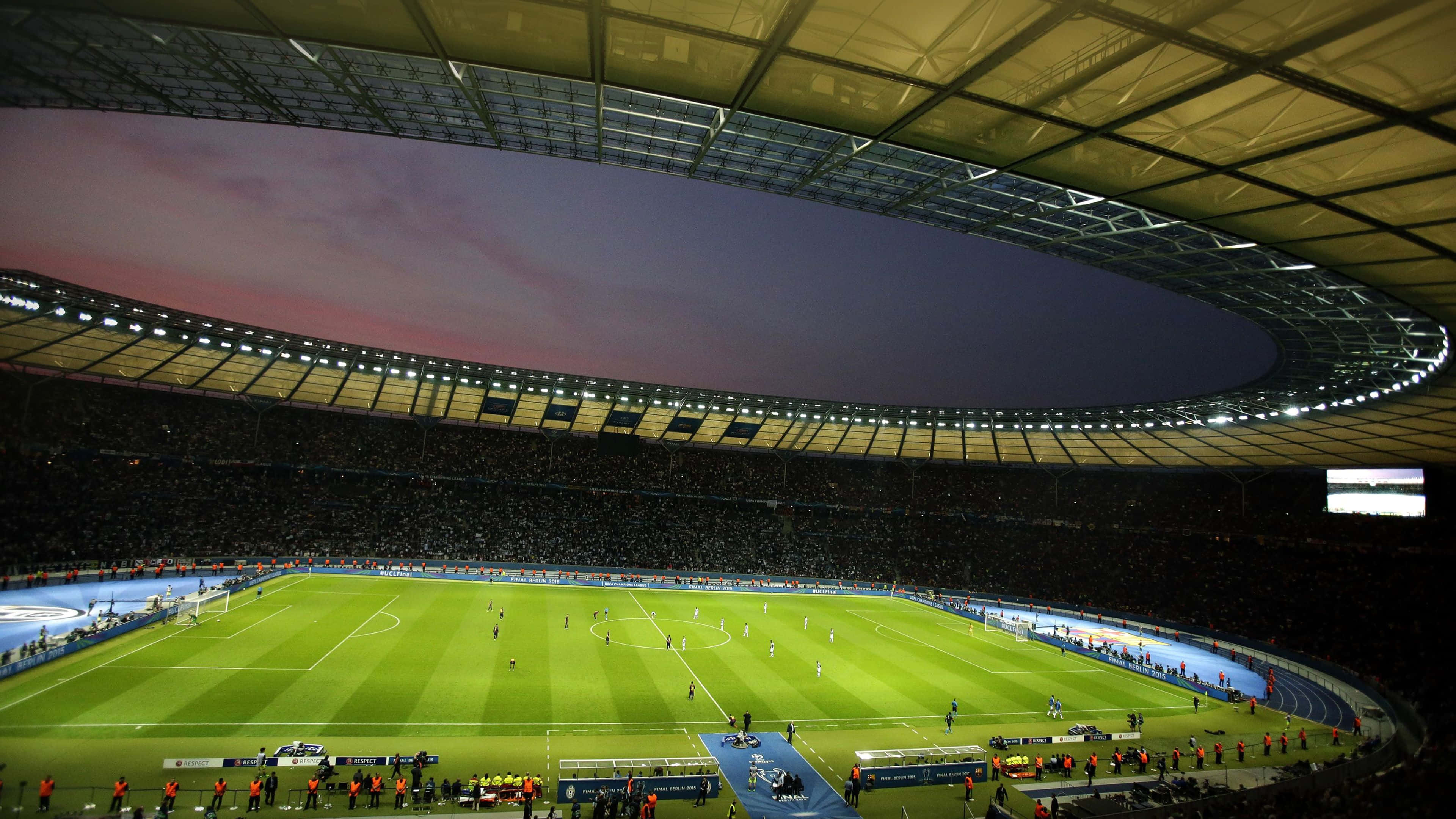 Soccer 4K 桌布 壁紙 — 黃昏時分，一個寬闊的照明足球場，翠綠的草坪和擠滿觀眾的看台，在暮色天空下。