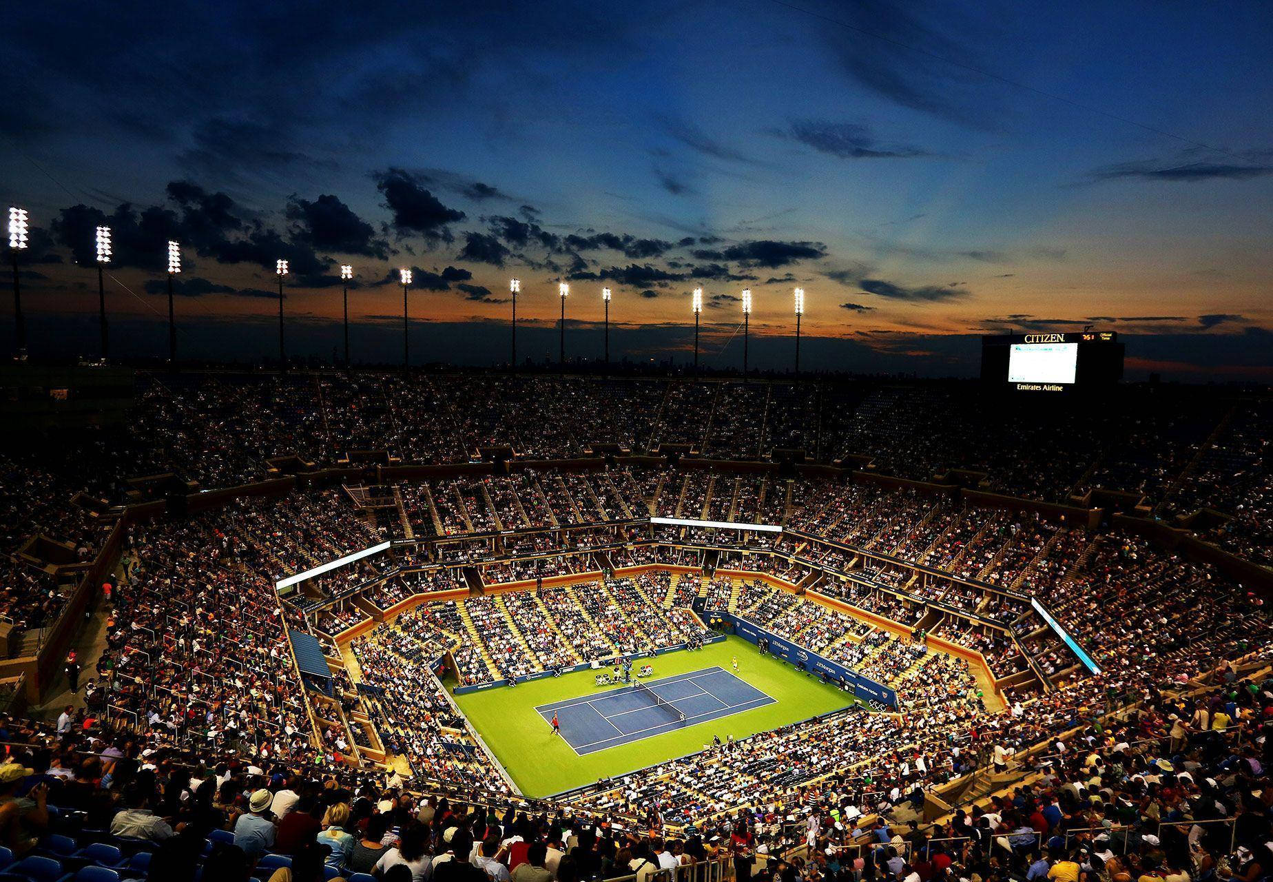 Tennis 桌布 壁紙 — 黃昏時分，一個座無虛席的網球場，擁有充滿活力的藍色球場和閃爍的泛光燈，映襯著暮色天空。