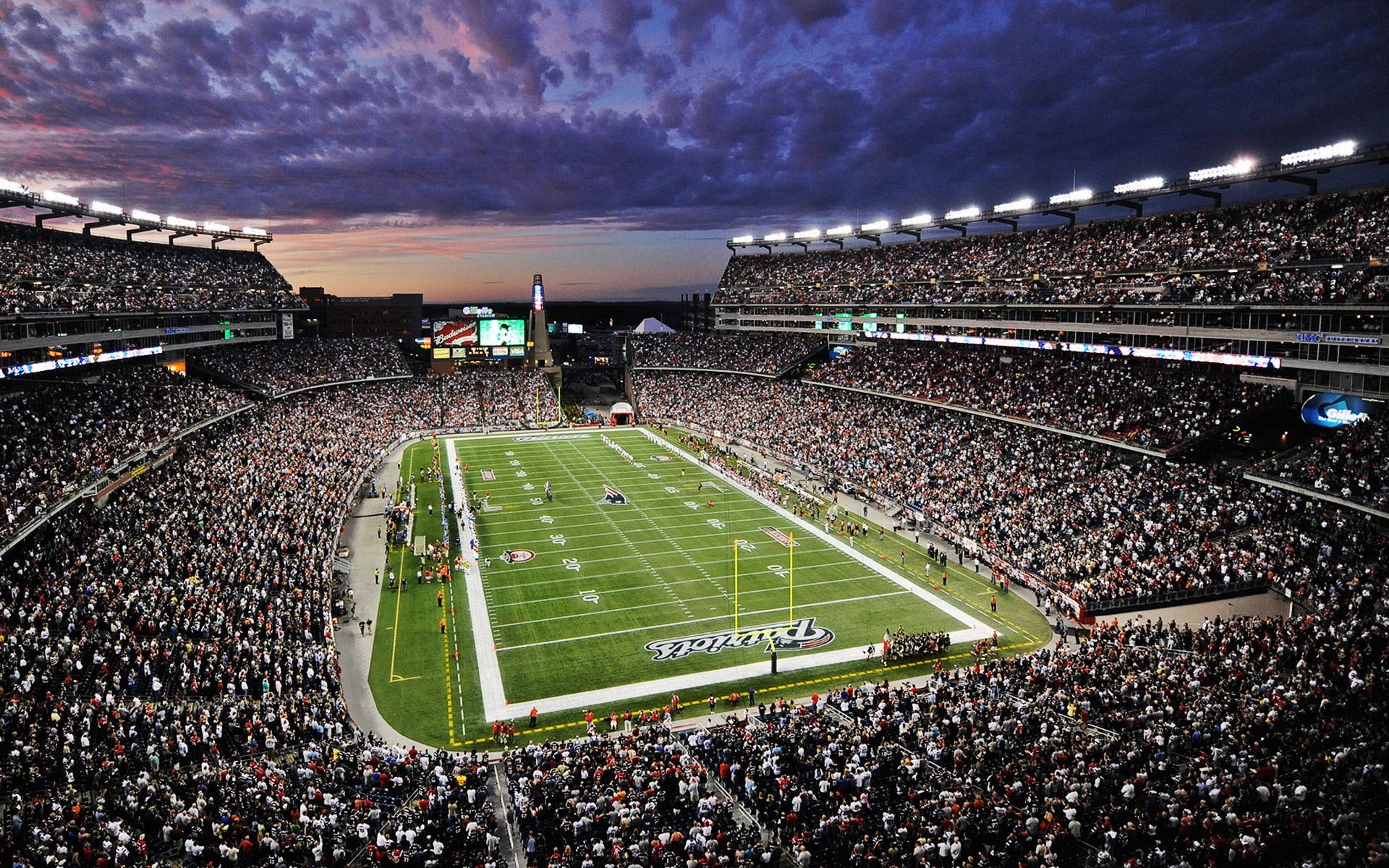 Stadium 桌布 壁紙 — 黃昏時分，一個擠滿人的足球場的鳥瞰圖，充滿活力的紫色雲彩和發光的場地燈光照亮人群。