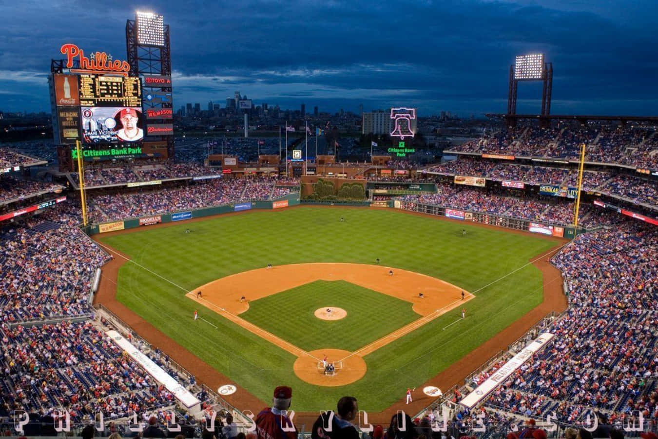 Baseball Field 桌布 壁紙 — 黃昏時分，一個擠滿觀眾的棒球場，翠綠的場地和發光的記分板在深藍色的天空下。