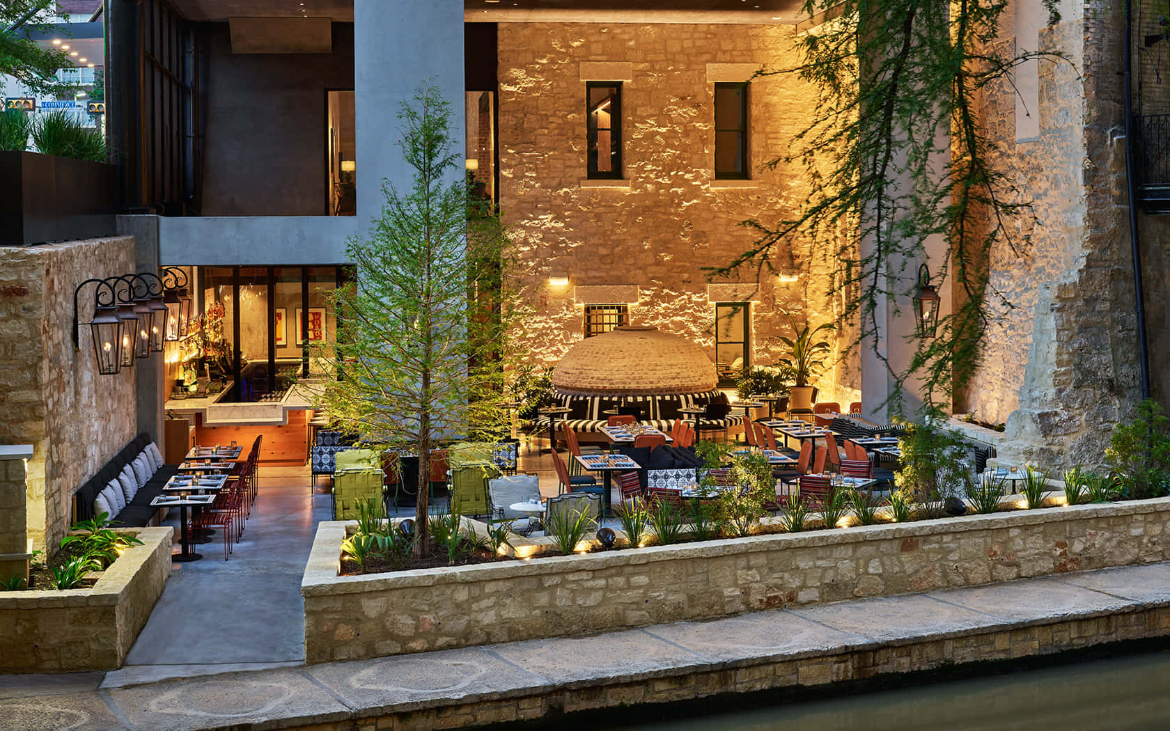 San Antonio 桌布 壁紙 — 黃昏時分，一個溫暖照明的戶外餐廳庭院，有石牆、綠植和桌子，旁邊是一條平靜的水道。