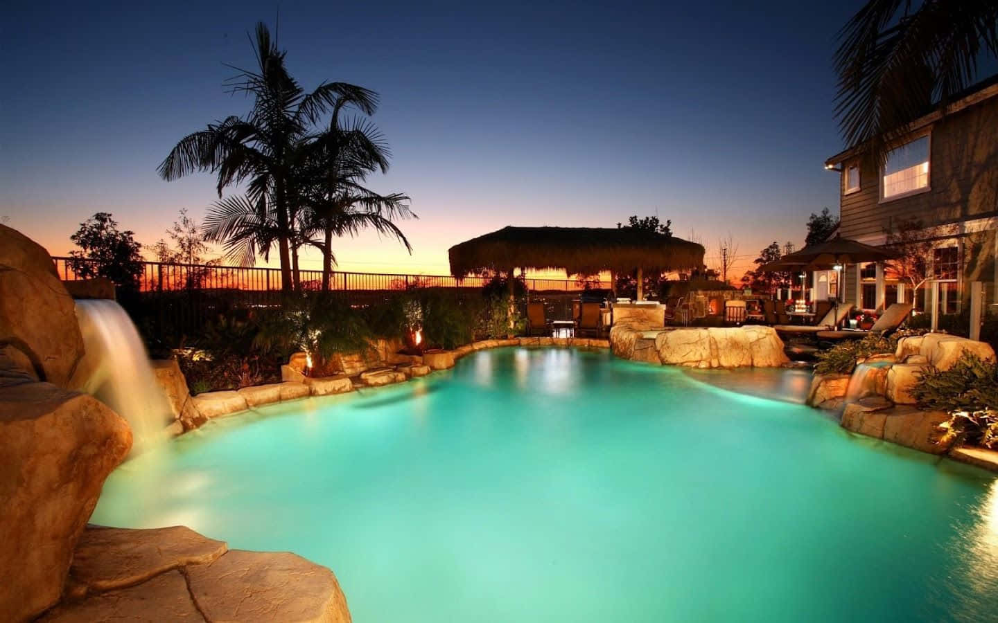 Resort 桌布 壁紙 — 黃昏時分，一個發光的藍綠色泳池，周圍環繞著棕櫚樹和石頭元素，帶有溫暖的環境照明。