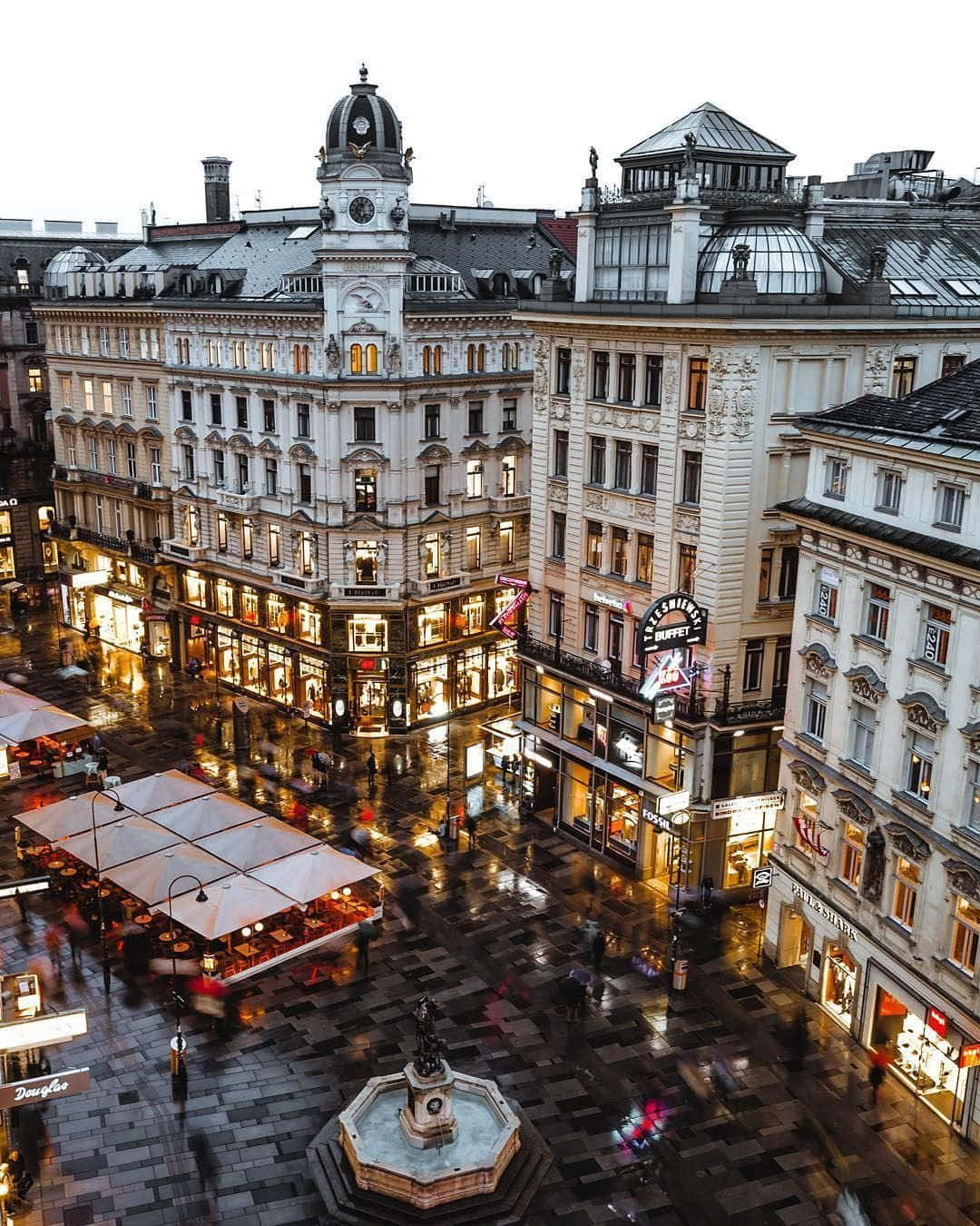 Europe 桌布 壁紙 — 黃昏時分，一個被雨水打濕的歐洲城市廣場，閃爍的商店櫥窗和中央噴泉反射著溫暖的燈光。